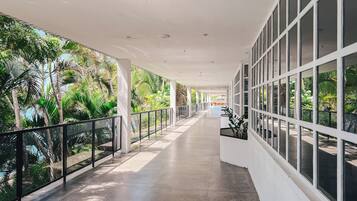 Um corredor coberto com piso ladrilhado, grandes janelas e vista para a vegetação tropical.