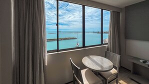 A room with a large window offering a view of a calm sea and a pier. There is a round table and two chairs in the room. - Marina Park (Fortaleza)