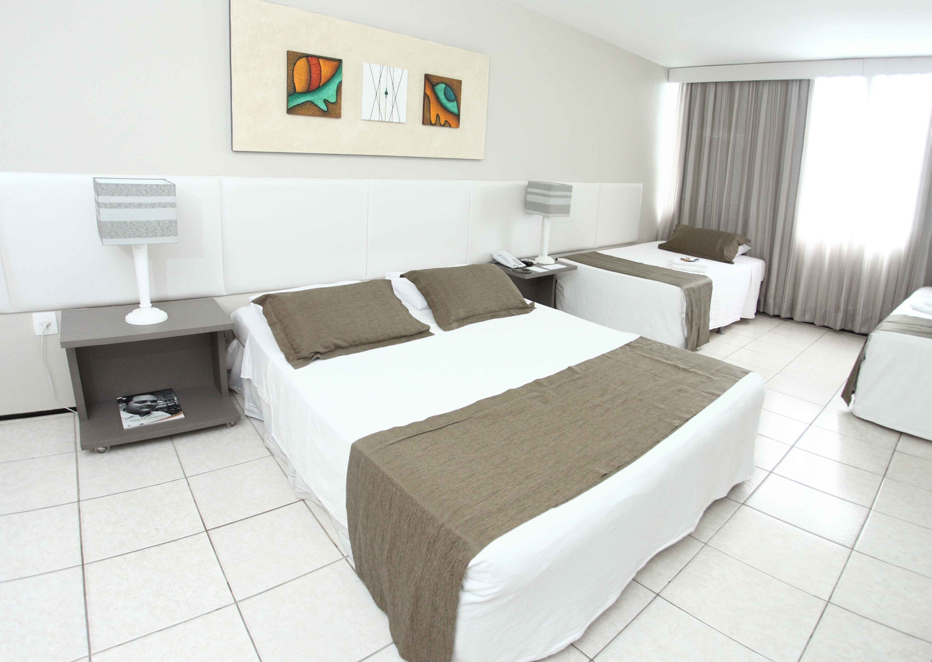 A hotel room with a large bed, bedside tables, a desk, and a chair. There are three paintings on the wall.