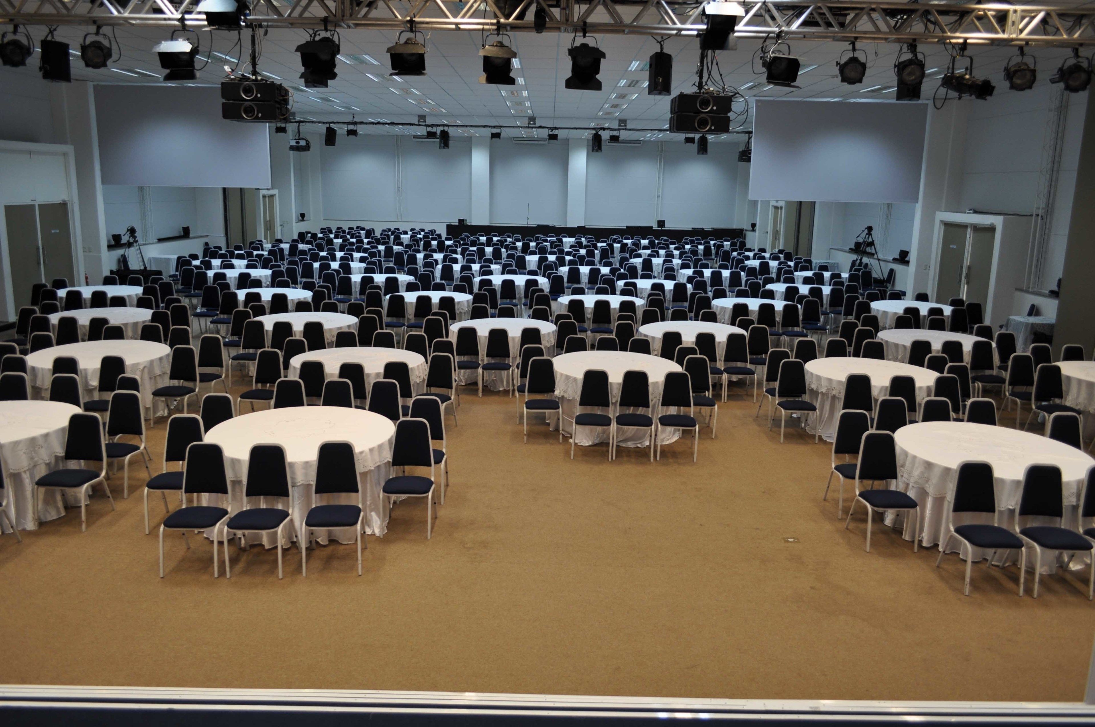 Uma grande sala de conferências com várias mesas redondas e cadeiras, equipada com iluminação no teto e uma tela grande na frente.