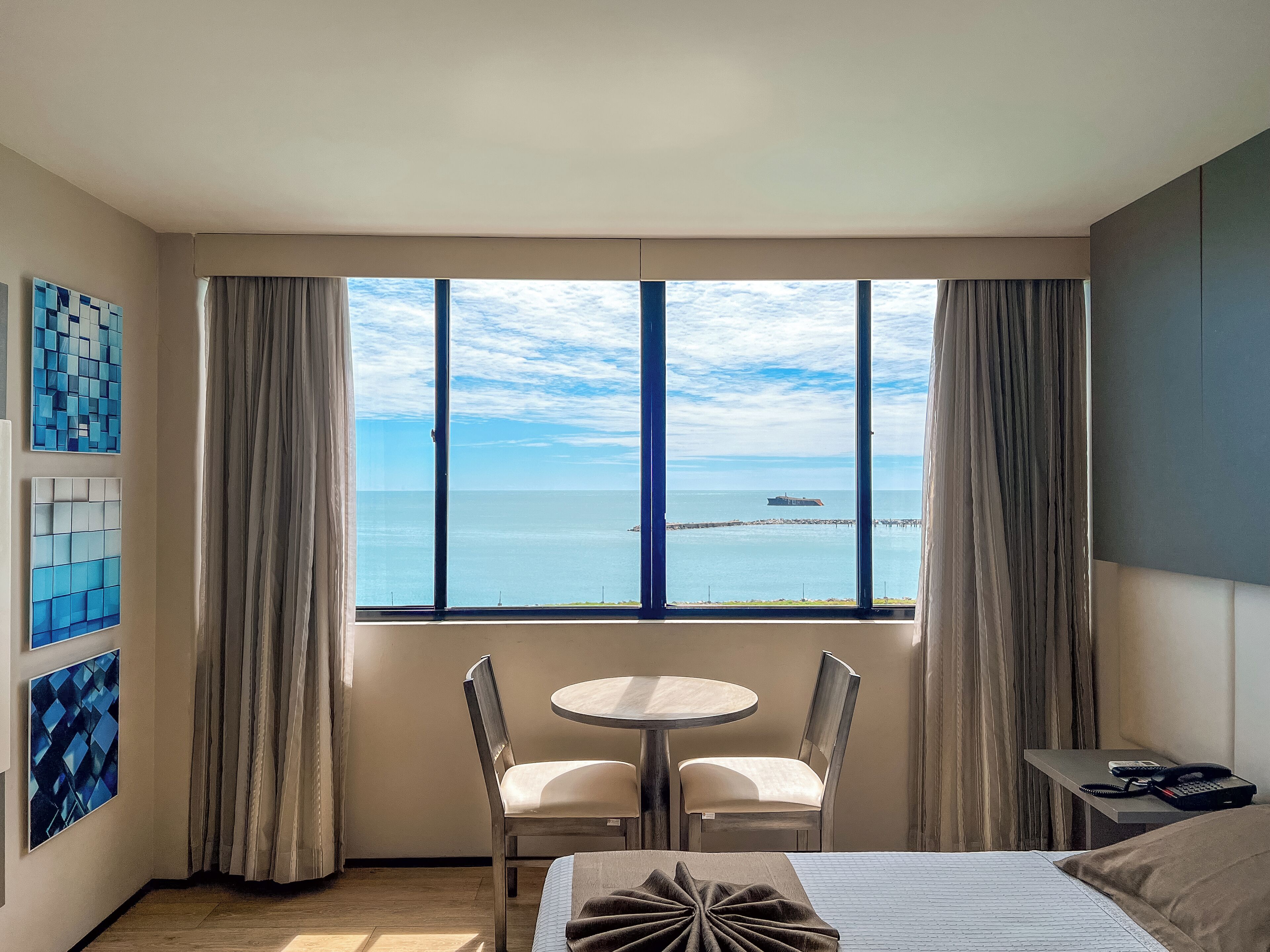 A hotel room with a large window offering a view of the sea and a pier. There is a bed, a small round table with two chairs, and a nightstand with a phone and remote control.