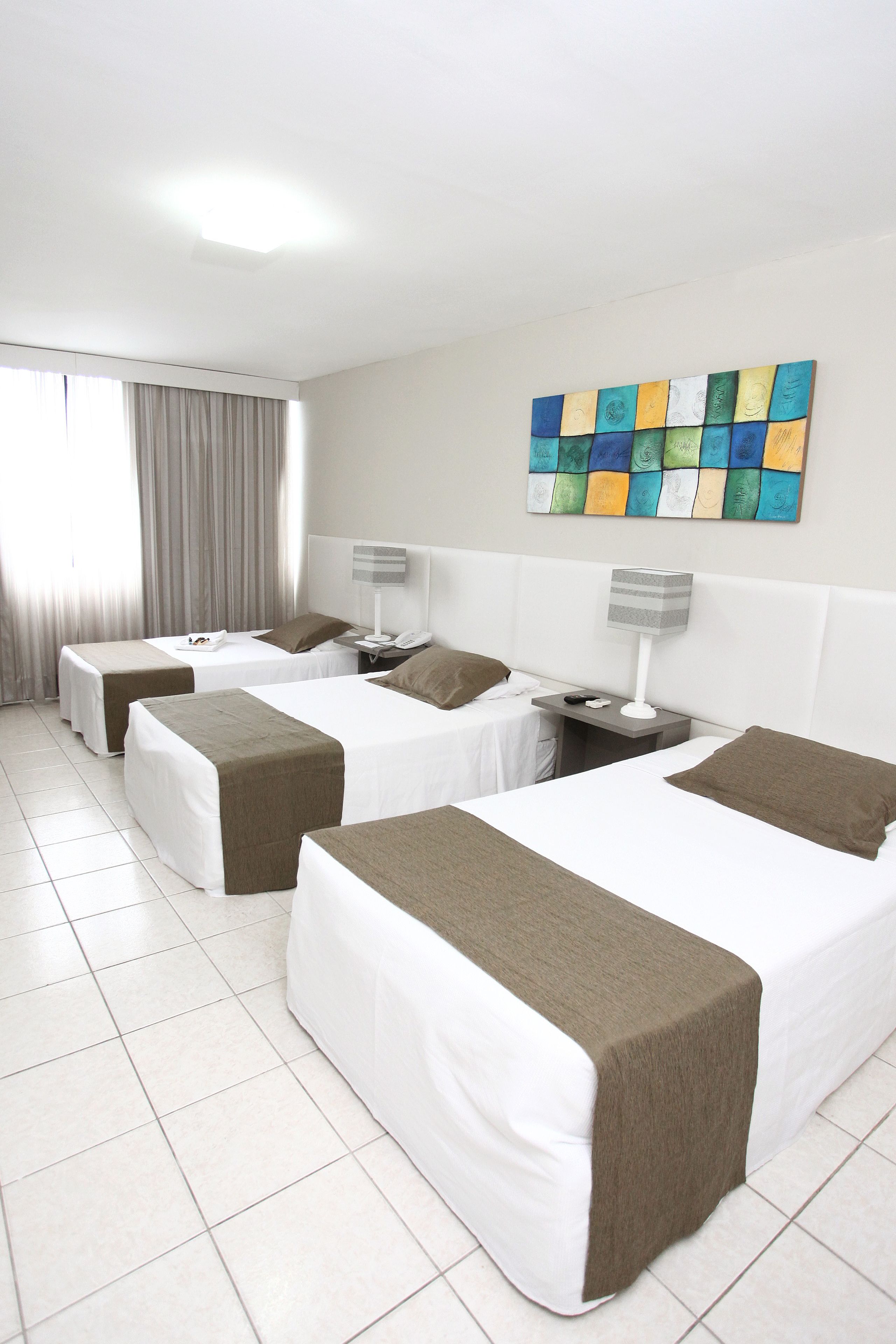 Quarto de hotel com três camas, uma janela grande com cortinas, uma obra de arte na parede e piso ladrilhado.
