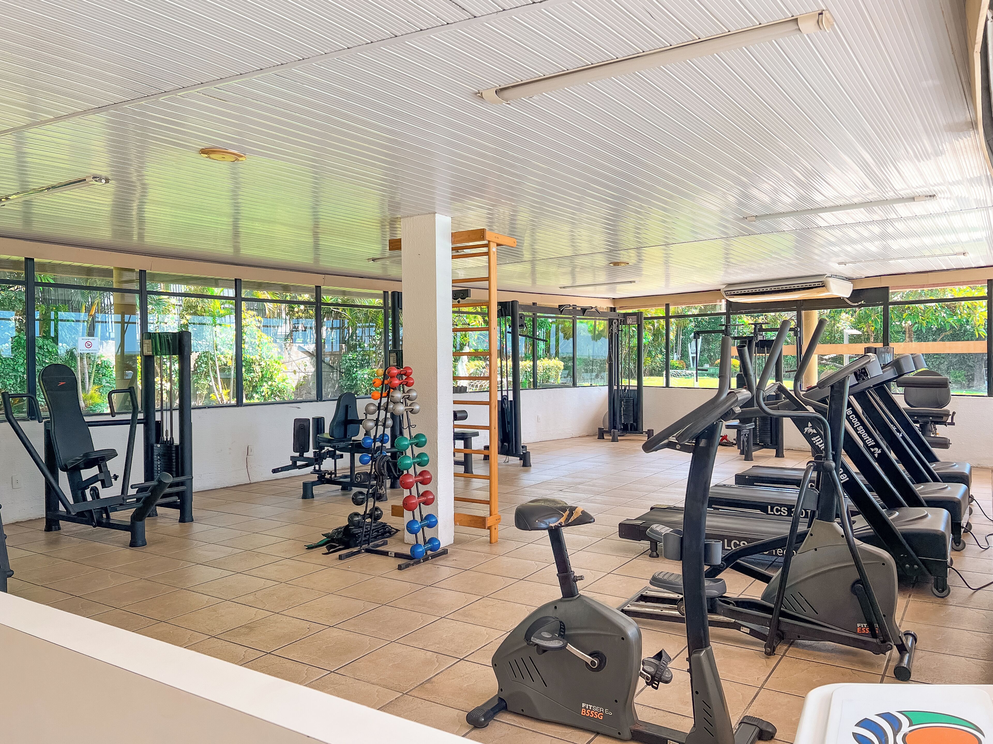 A spacious gym with various exercise equipment, large windows, and a tiled floor.