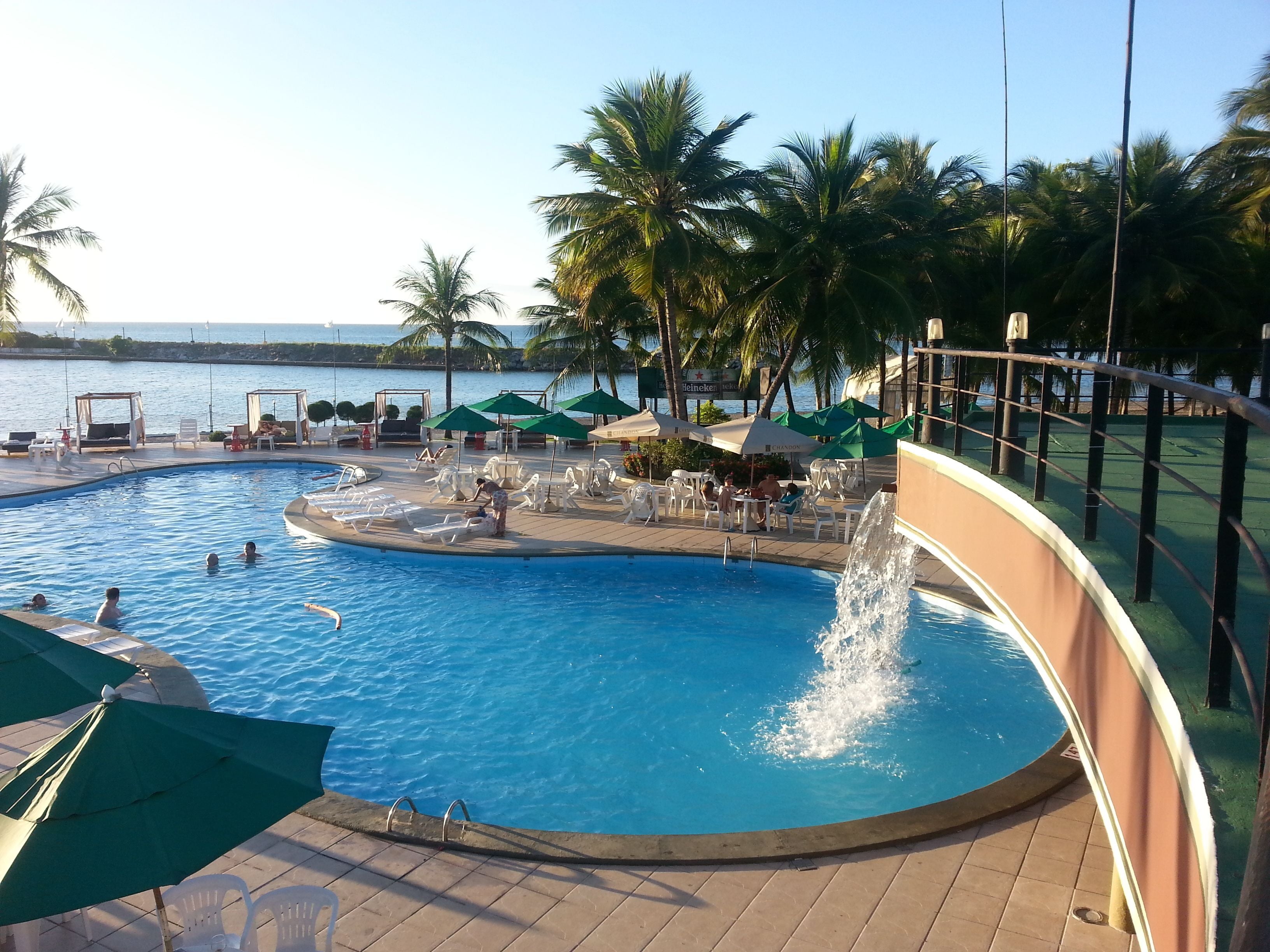 Piscina com uma fonte de água, rodeada por espreguiçadeiras e guarda-sóis, palmeiras e vista para o mar.