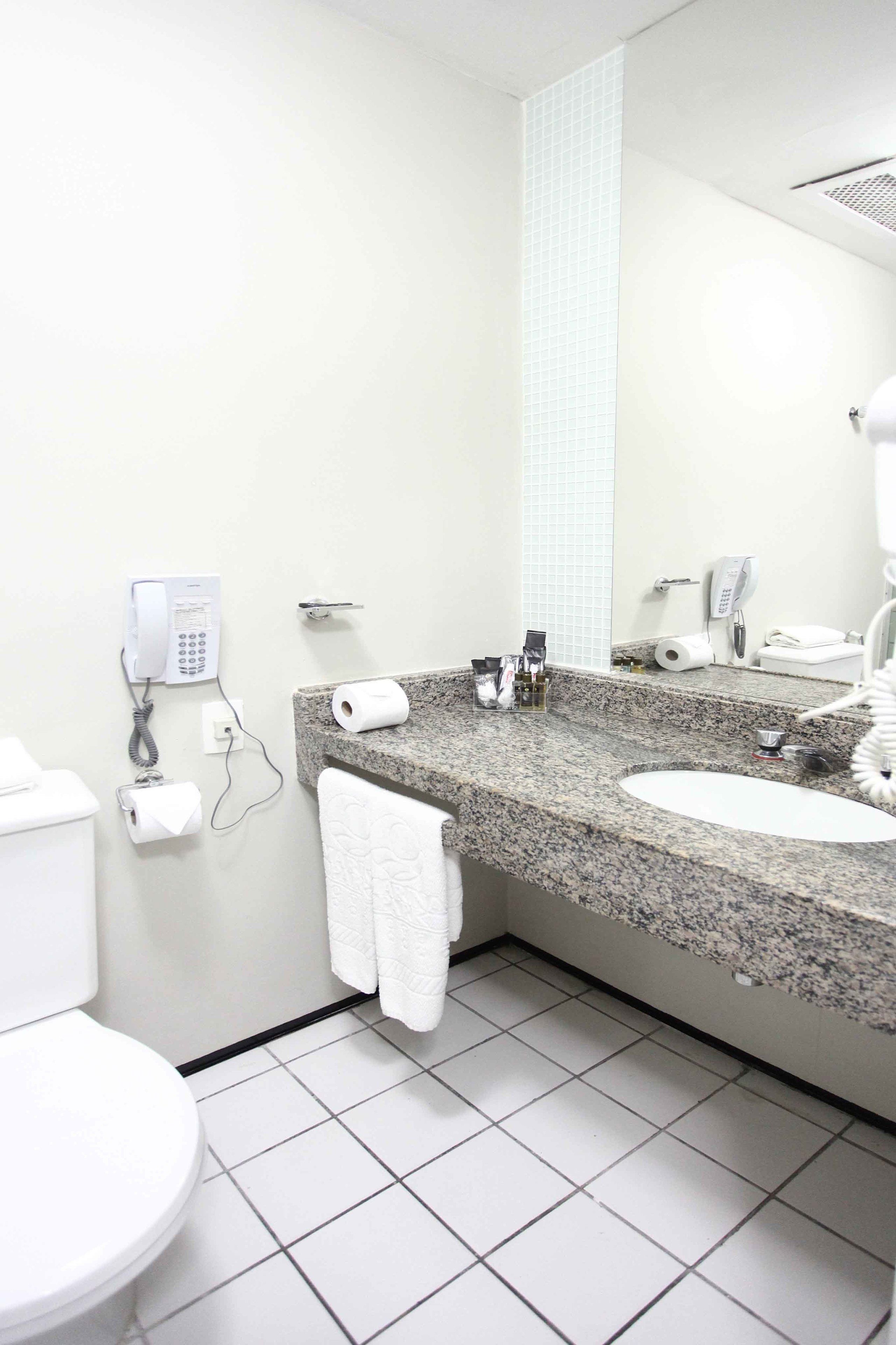 Un baño moderno con encimera de granito, lavabo blanco y un espejo grande. Hay dos teléfonos instalados en la pared, un perchero y un inodoro.