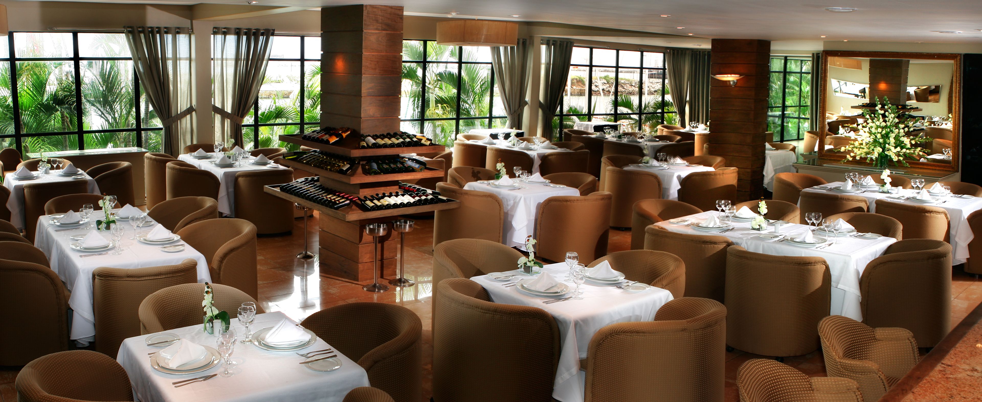 A restaurant with multiple tables set for dining, a wine rack, and large windows offering a view of greenery.