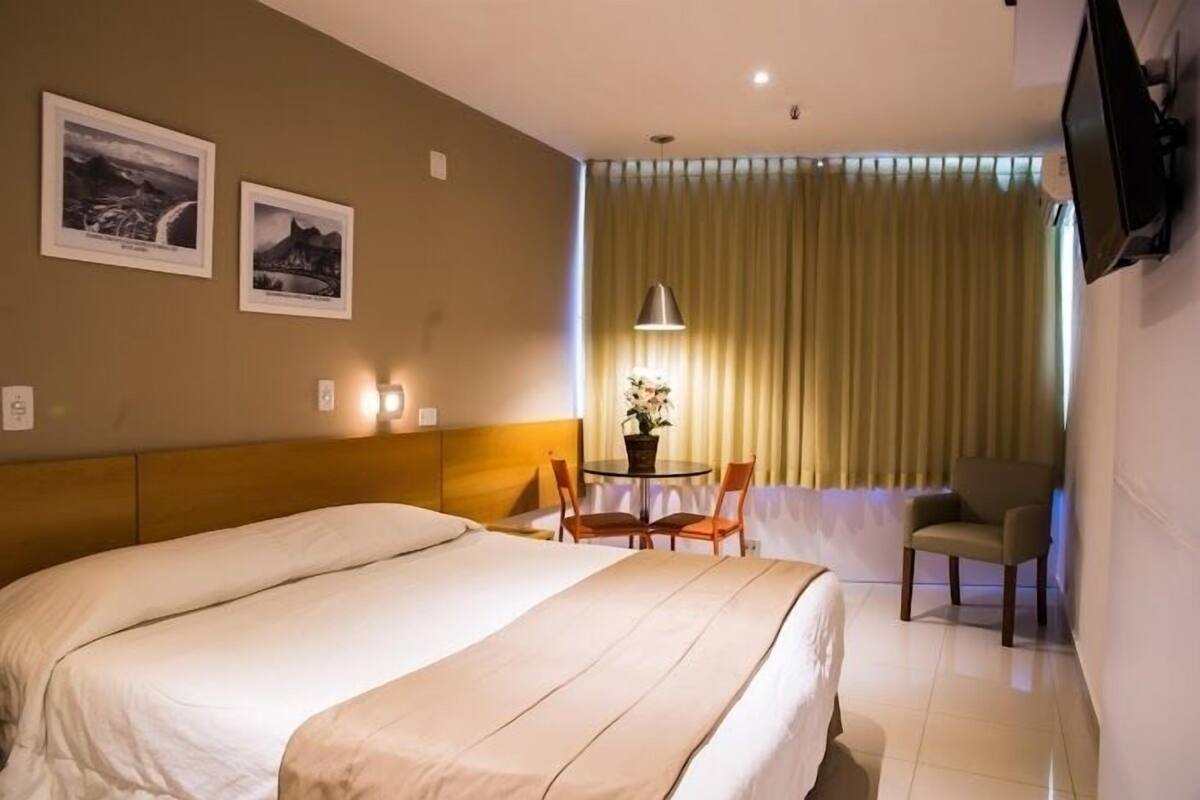 Ein Hotelzimmer mit einem großen Bett, einem Schreibtisch mit zwei Stühlen, einem an der Wand montierten Fernseher und zwei gerahmten Bildern.