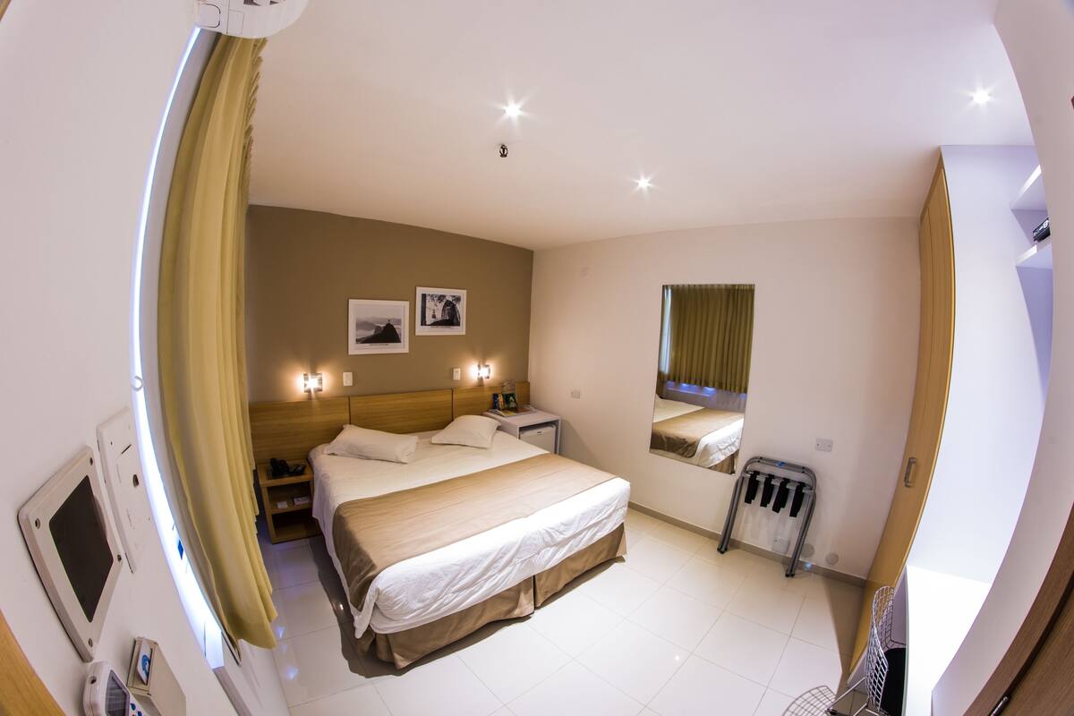 Ein Hotelzimmer mit Bett, Nachttisch, Lampe, Spiegel und einem Fenster mit Vorhängen.