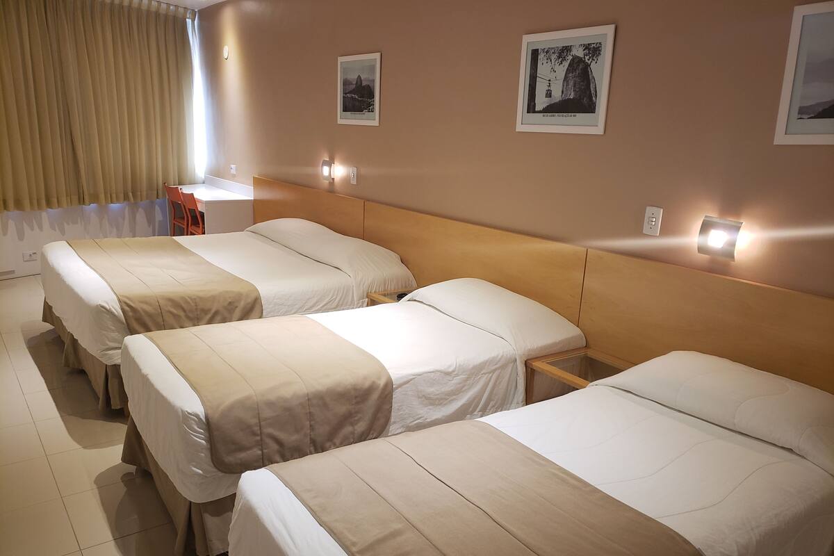 Ein Hotelzimmer mit drei Einzelbetten, einem Holzkopfende, einem kleinen Tisch und zwei Wandleuchten.
