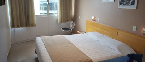 Habitación de hotel con cama, una silla, ventana con cortinas, equipo de aire acondicionado y dos cuadros en la pared.