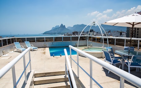 A rooftop pool area with a view of mountains, lounge chairs, and a table with umbrellas.