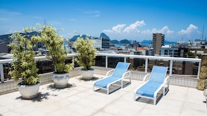 Una terraza en la azotea con sillones de descanso y plantas en macetas, con vistas a un paisaje urbano que incluye edificios y montañas.