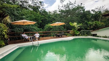 A swimming pool with two people sitting under umbrellas, surrounded by lush greenery.