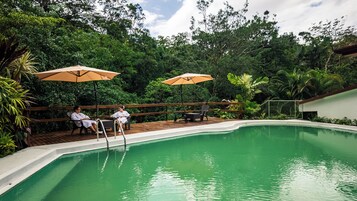 Una piscina con dos personas sentadas bajo sombrillas, rodeada de exuberante vegetación.