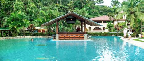 Une piscine dotée d’une terrasse en bois et d’un kiosque, entourée d’une végétation luxuriante et avec un bâtiment visible au loin.