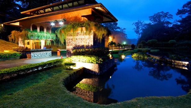 A well-lit building with a stone facade, a pond, and a landscaped garden at dusk. - Hotel El Tucano Resort & Thermal Spa (La Palmeria)