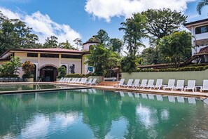 A swimming pool with lounge chairs, surrounded by greenery and a building with a red roof.