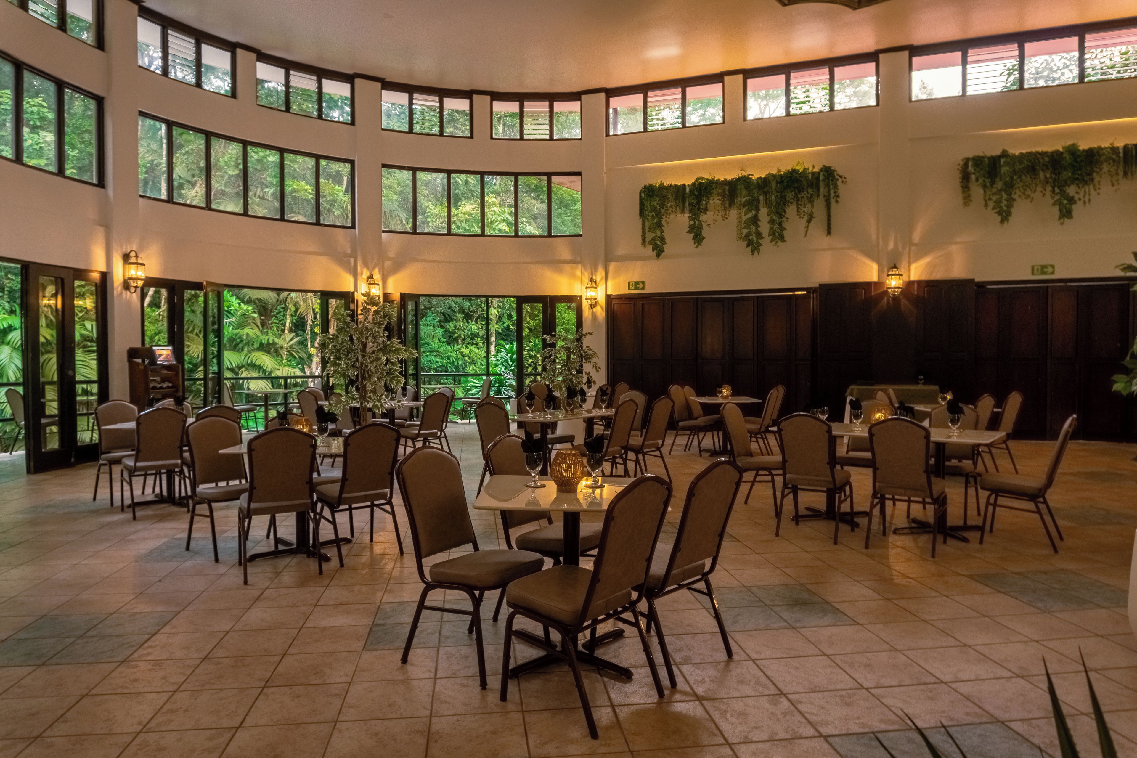 A spacious indoor dining area with multiple tables and chairs, large windows, and indoor plants.