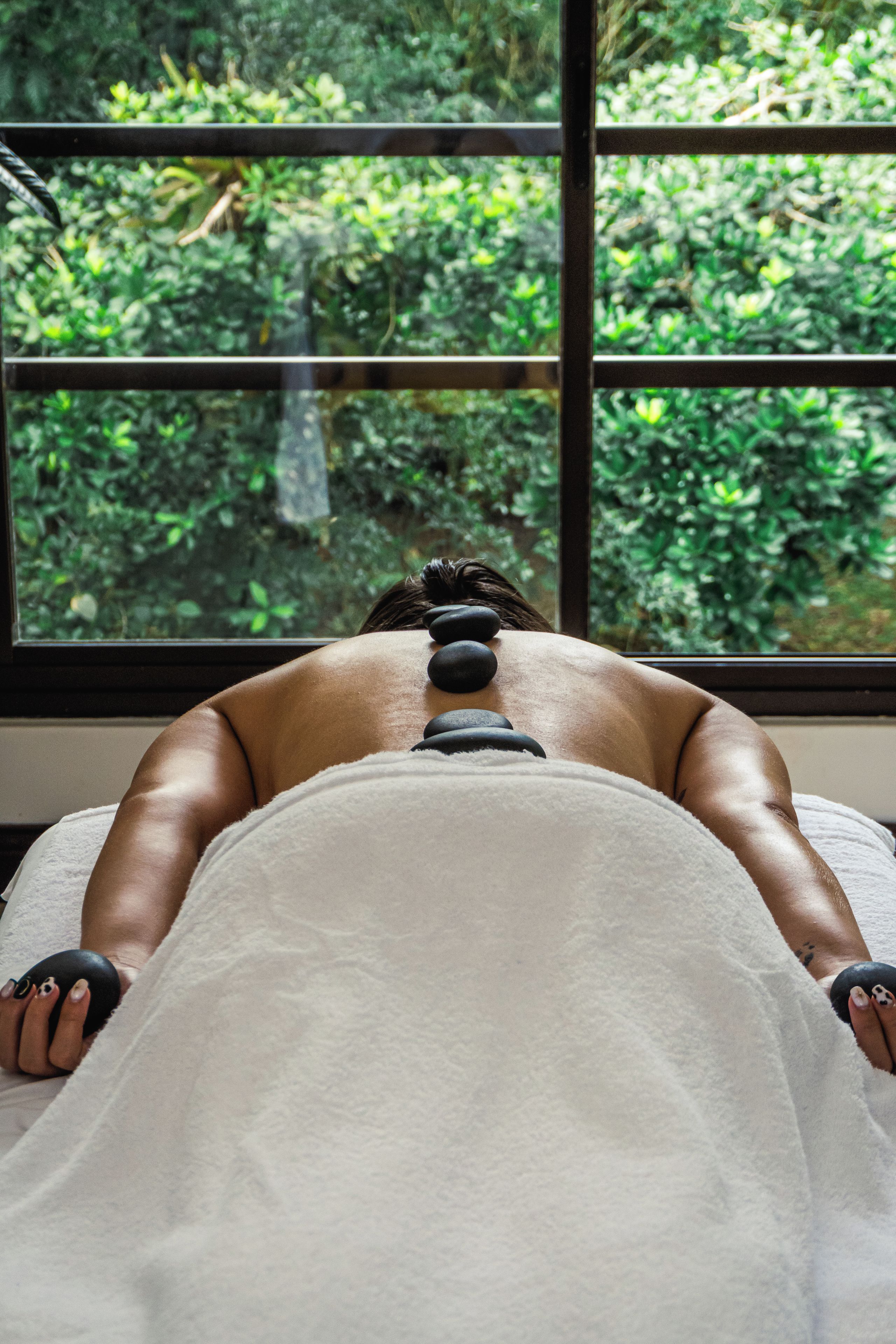 A person receiving a back massage with stones, with a window view of greenery.
