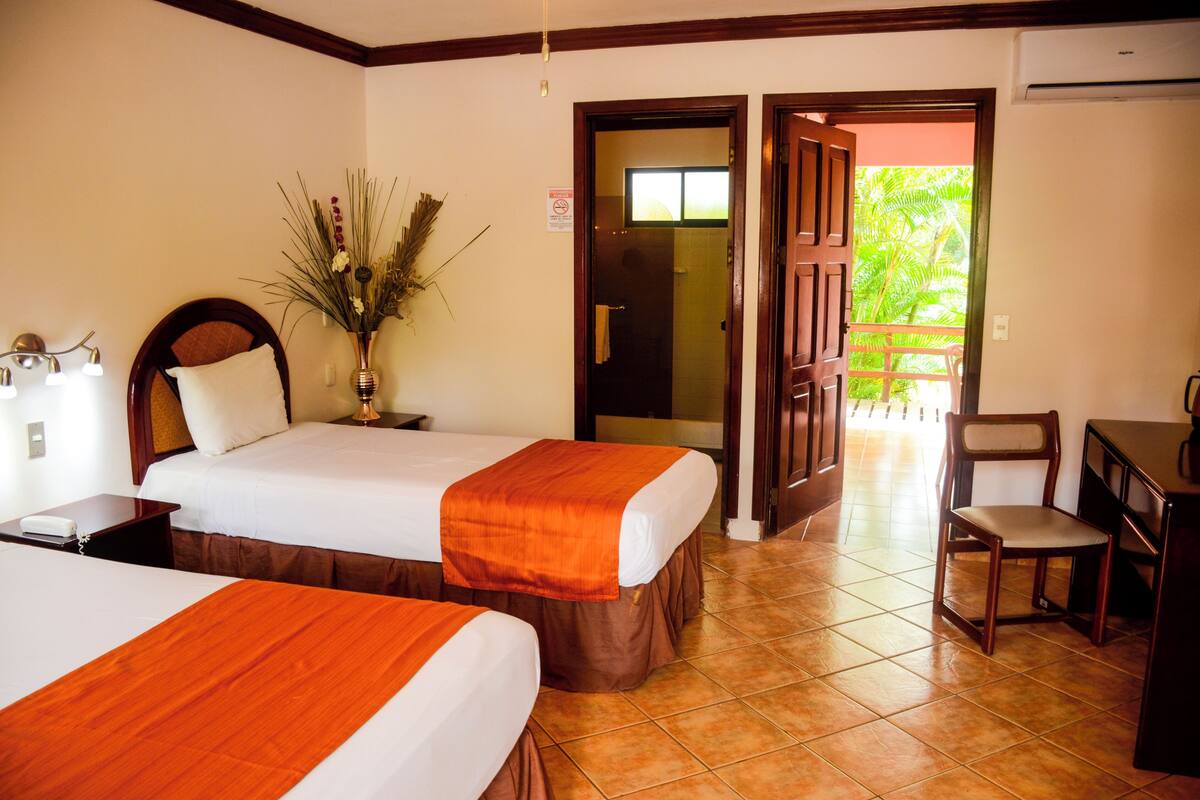 A hotel room with two beds, a desk, a chair, a vase with dried plants, and a door leading to a balcony.