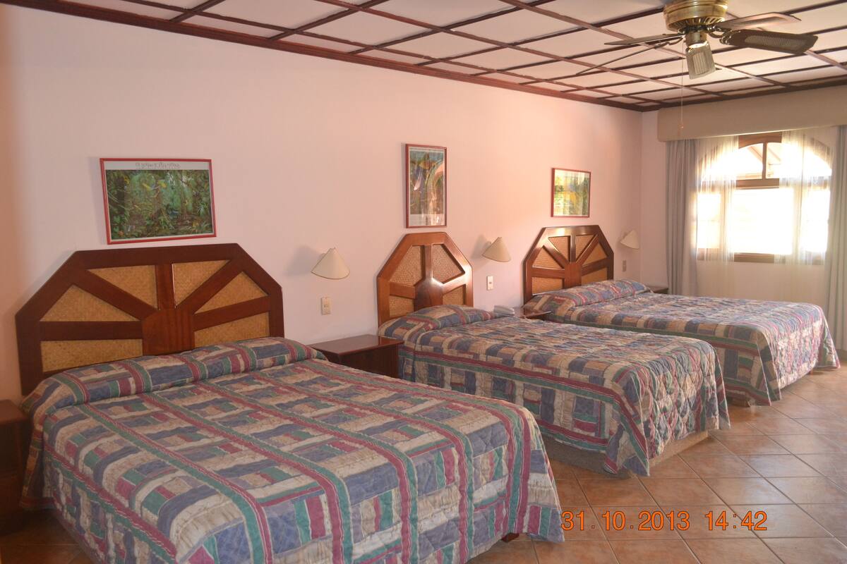 A hotel room with three beds, wooden headboards, and two wall-mounted lamps.