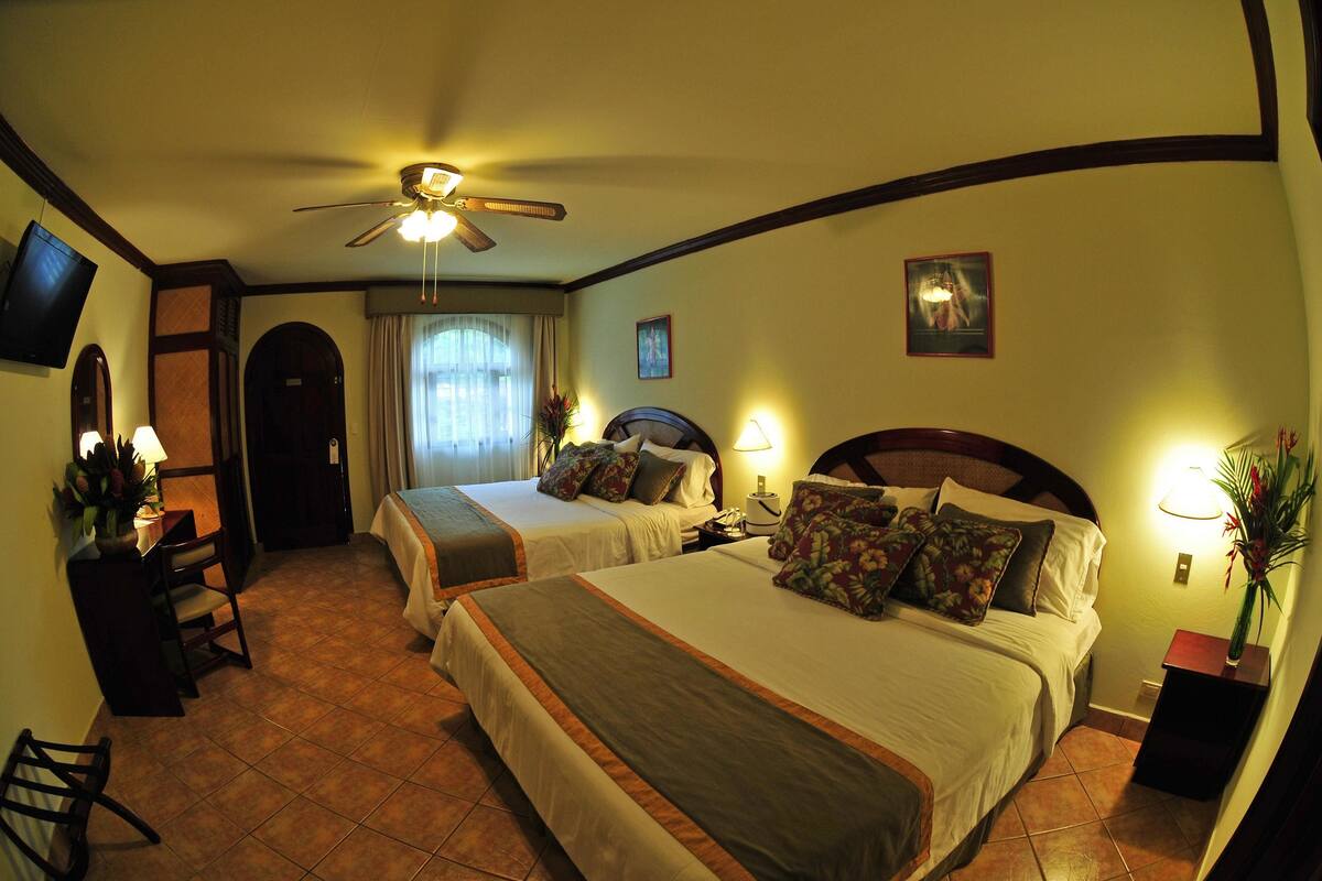 A hotel room with two beds, a desk, a chair, a television, and a ceiling fan.