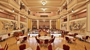 A spacious banquet hall with round tables and red chairs, featuring large wall paintings and a central buffet area. - Hotel Ariston & Ariston Patio (Prague)