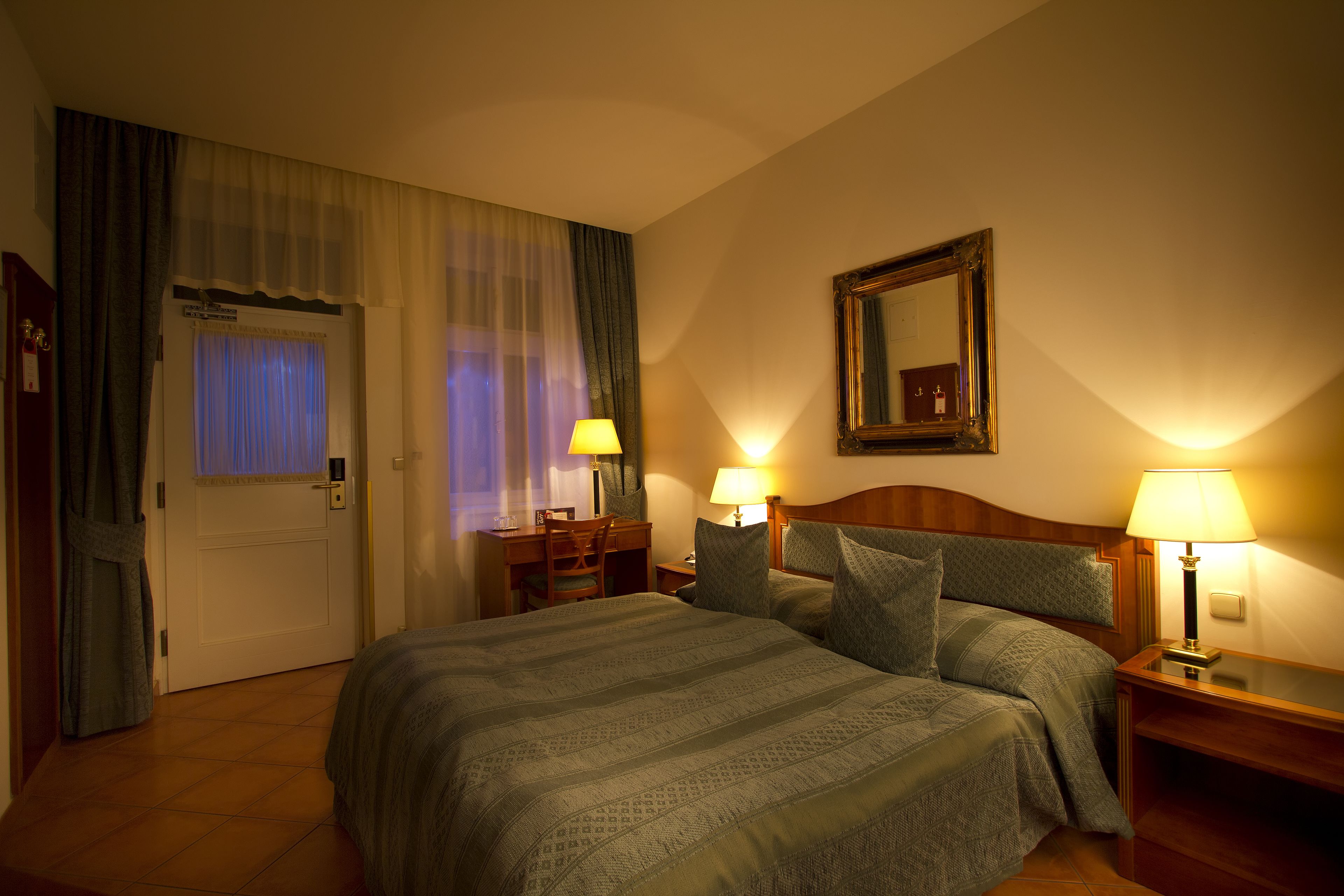 A hotel room with a large bed, two bedside tables with lamps, a wooden headboard, a desk with a chair, a window with curtains, and a mirror on the wall.