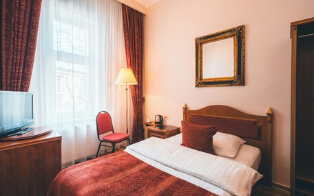 A hotel room with a bed, a desk with a television, a chair, a nightstand with a lamp, and a mirror on the wall.