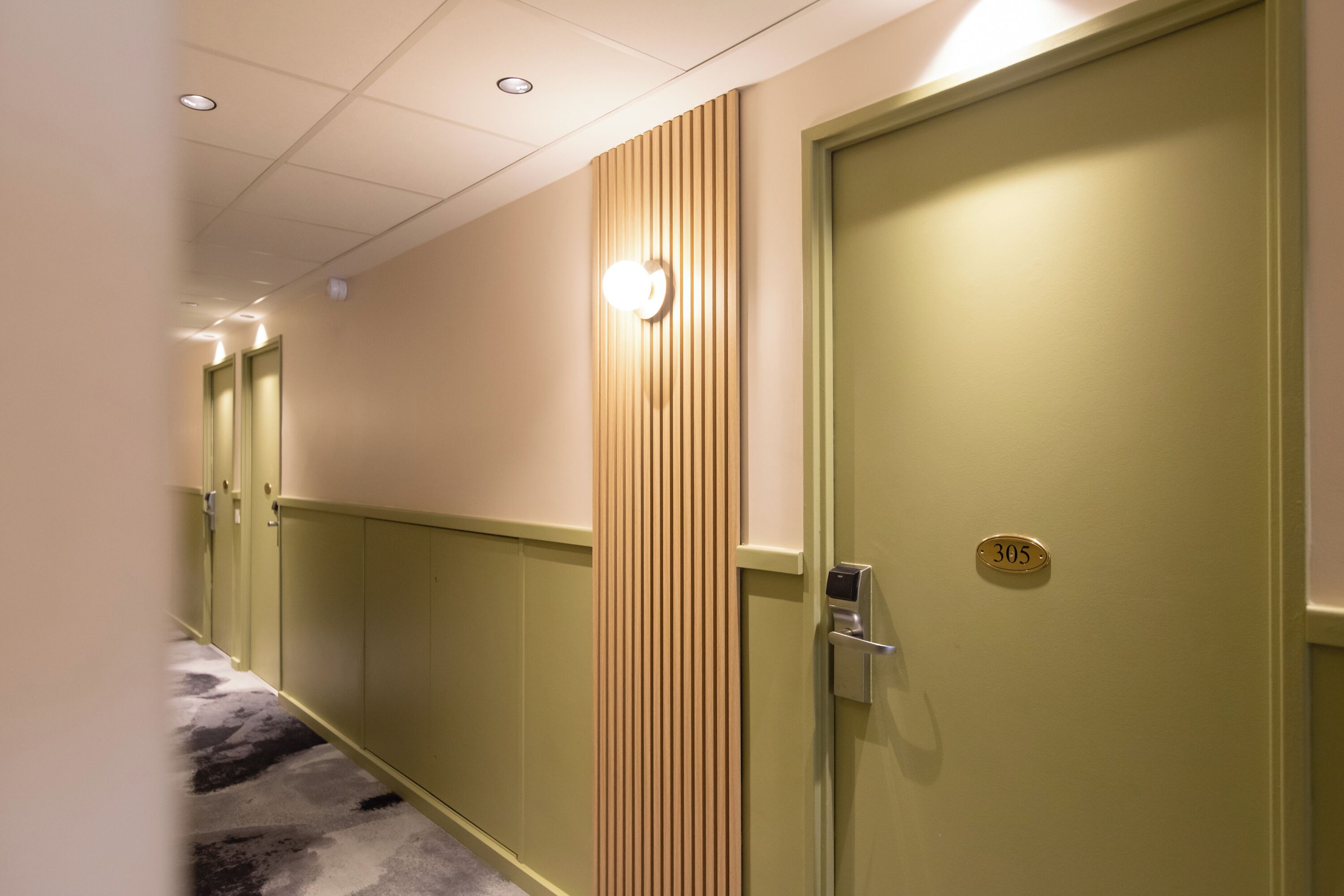 A hallway with a carpeted floor, a wooden panel wall, and a door with a number plate.