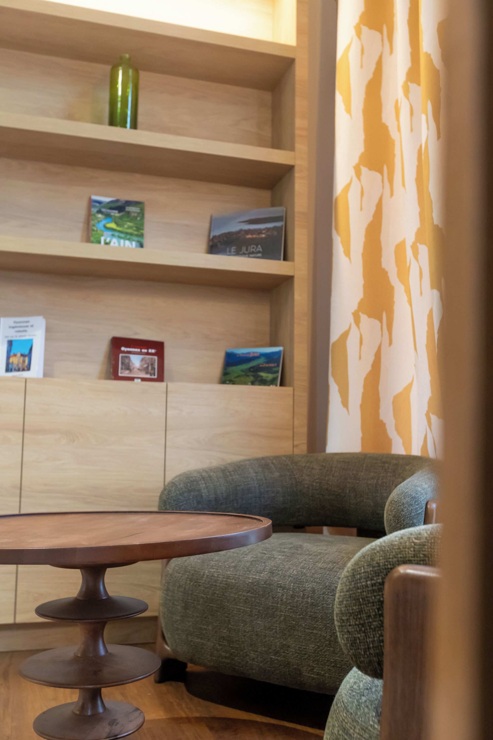 Ein Zimmer mit einem Holzbücherregal, einem grünen Polsterstuhl und einem runden Holztisch.