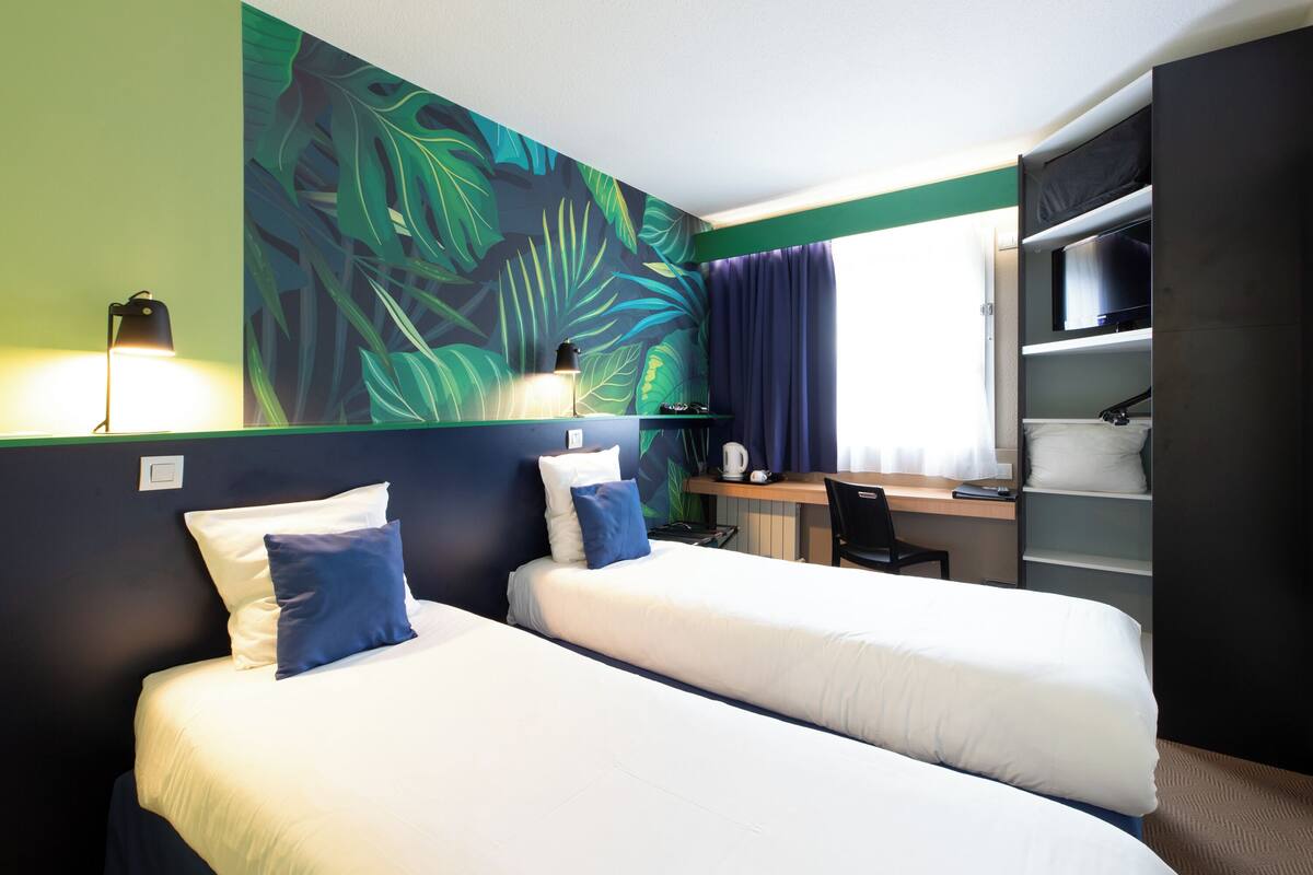 A hotel room with two beds, a desk, a chair, a TV, and a large window with curtains.