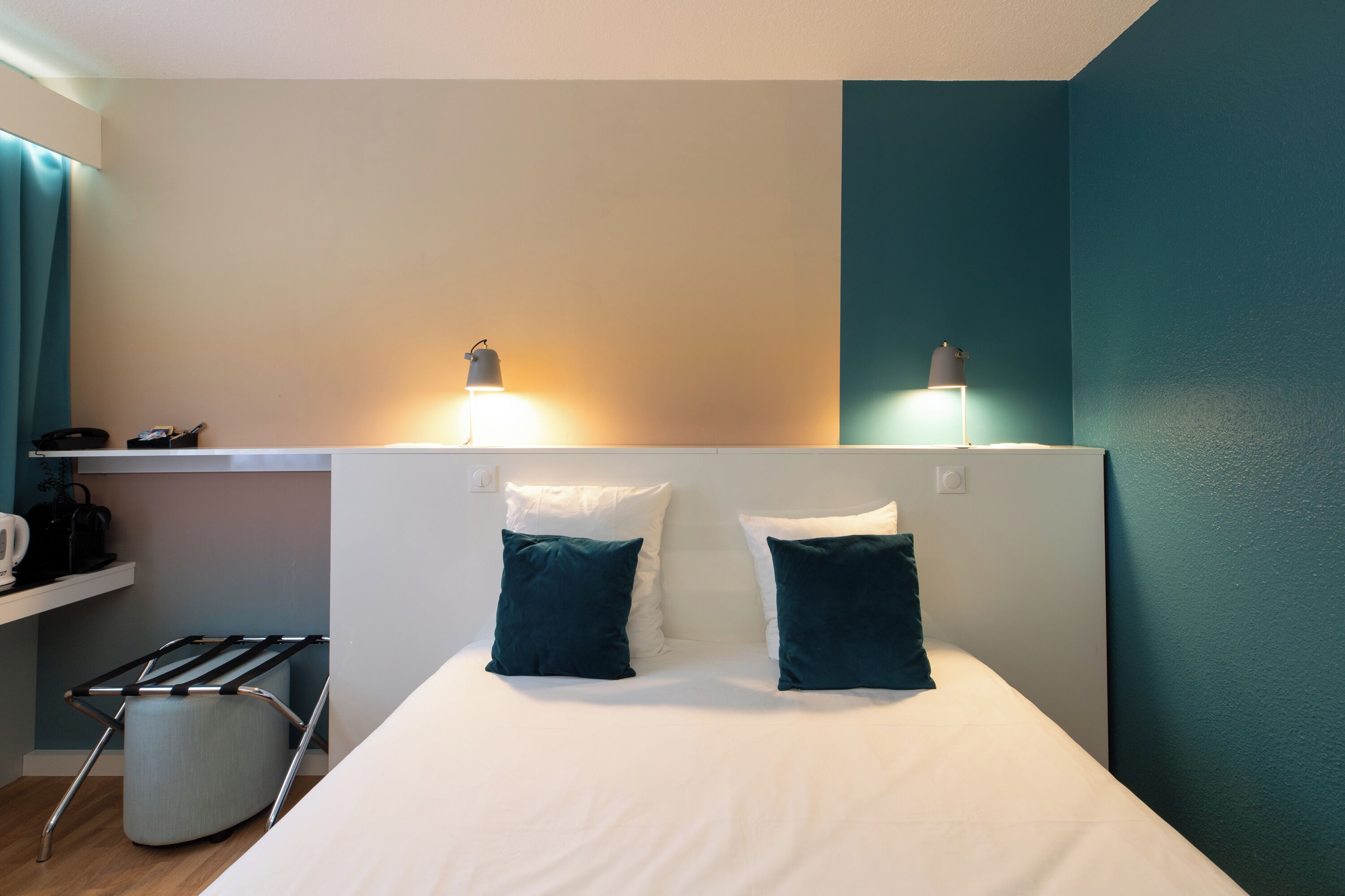 Una habitación de hotel con una cama, dos lámparas de noche, una mesita con lámpara y un estante en la pared.