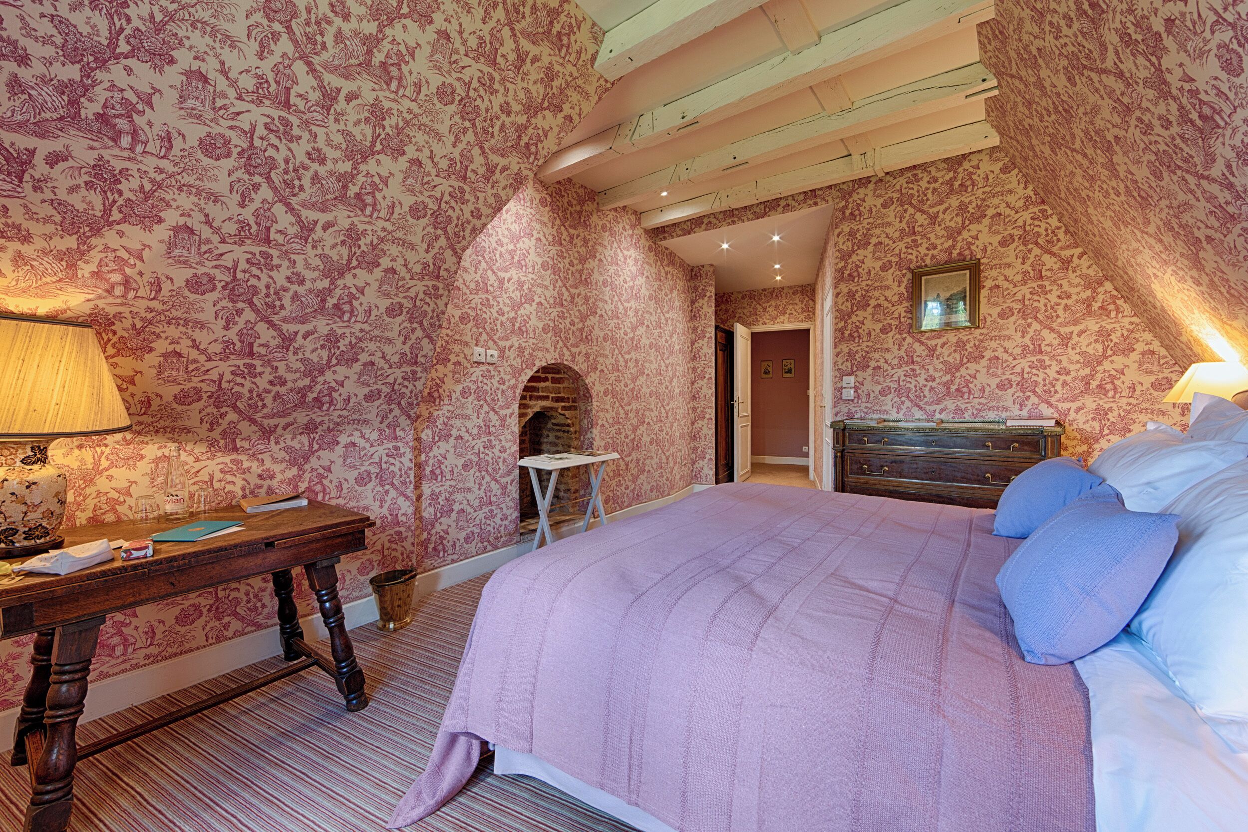 Une chambre avec un papier peint à motifs floraux, un lit en bois, une table de chevet, une lampe et une petite table.