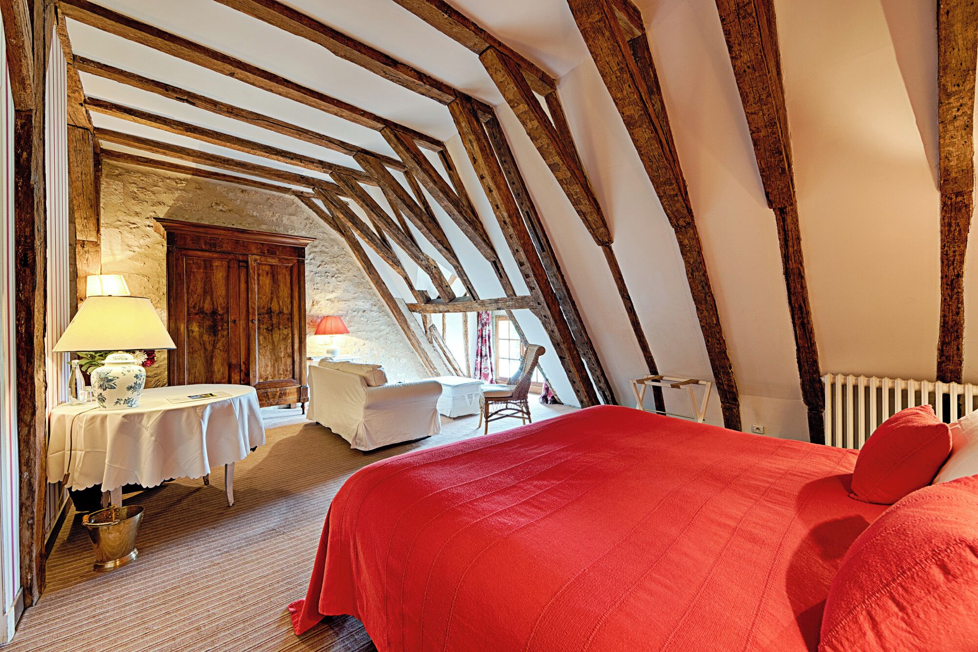 A bedroom with exposed wooden beams, a red bedspread, a white chair, and a table with a lamp.