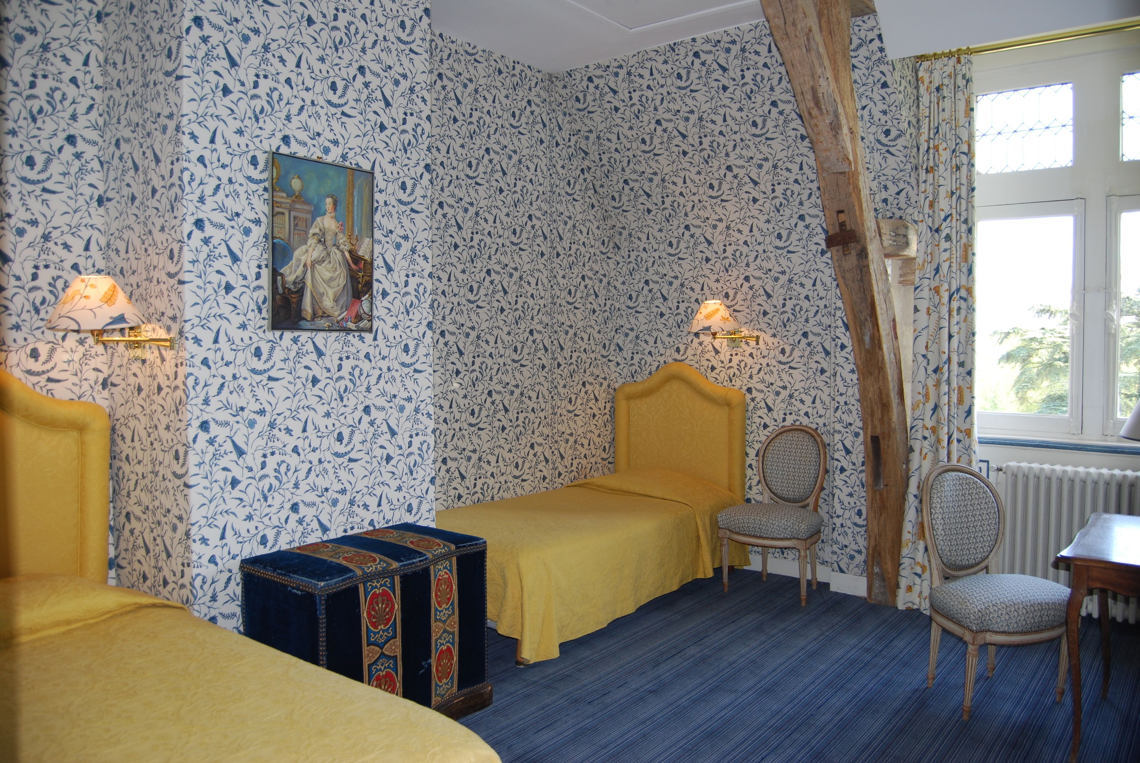 A room with patterned wallpaper, a bed with a yellow headboard, a small table, two chairs, and a window with curtains.