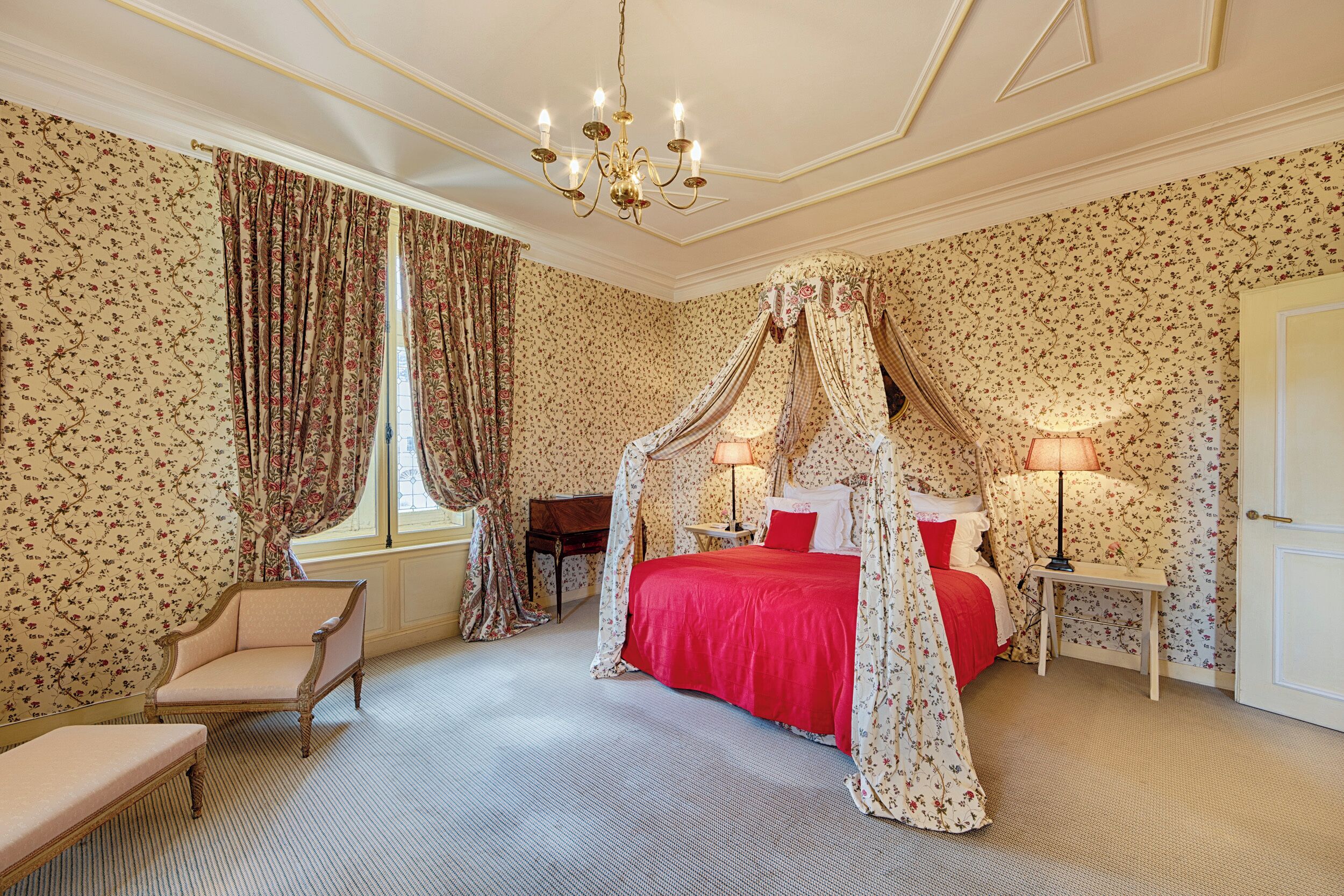A bedroom with a four-poster bed, a sitting chair, a bedside table, and a window with curtains.