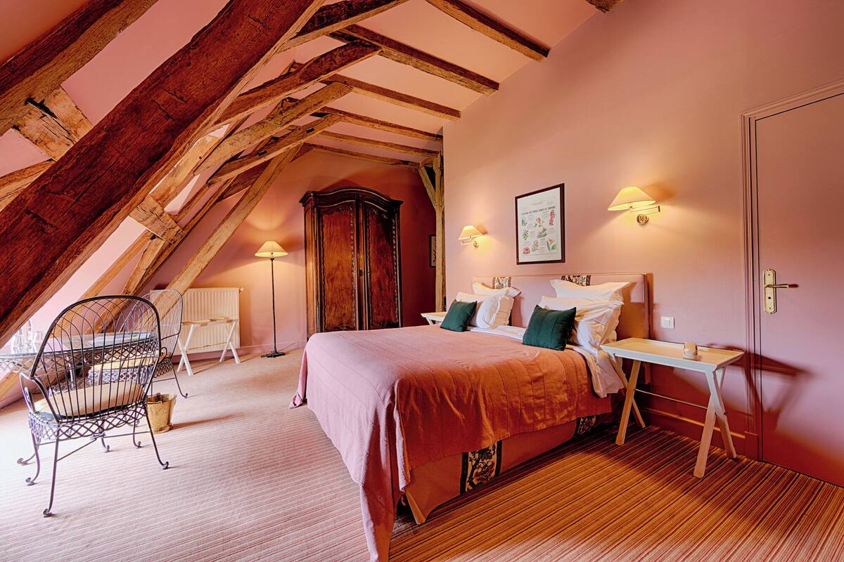 A cozy bedroom with a sloped ceiling, a bed with green pillows, a wooden wardrobe, and a small table with a lamp.