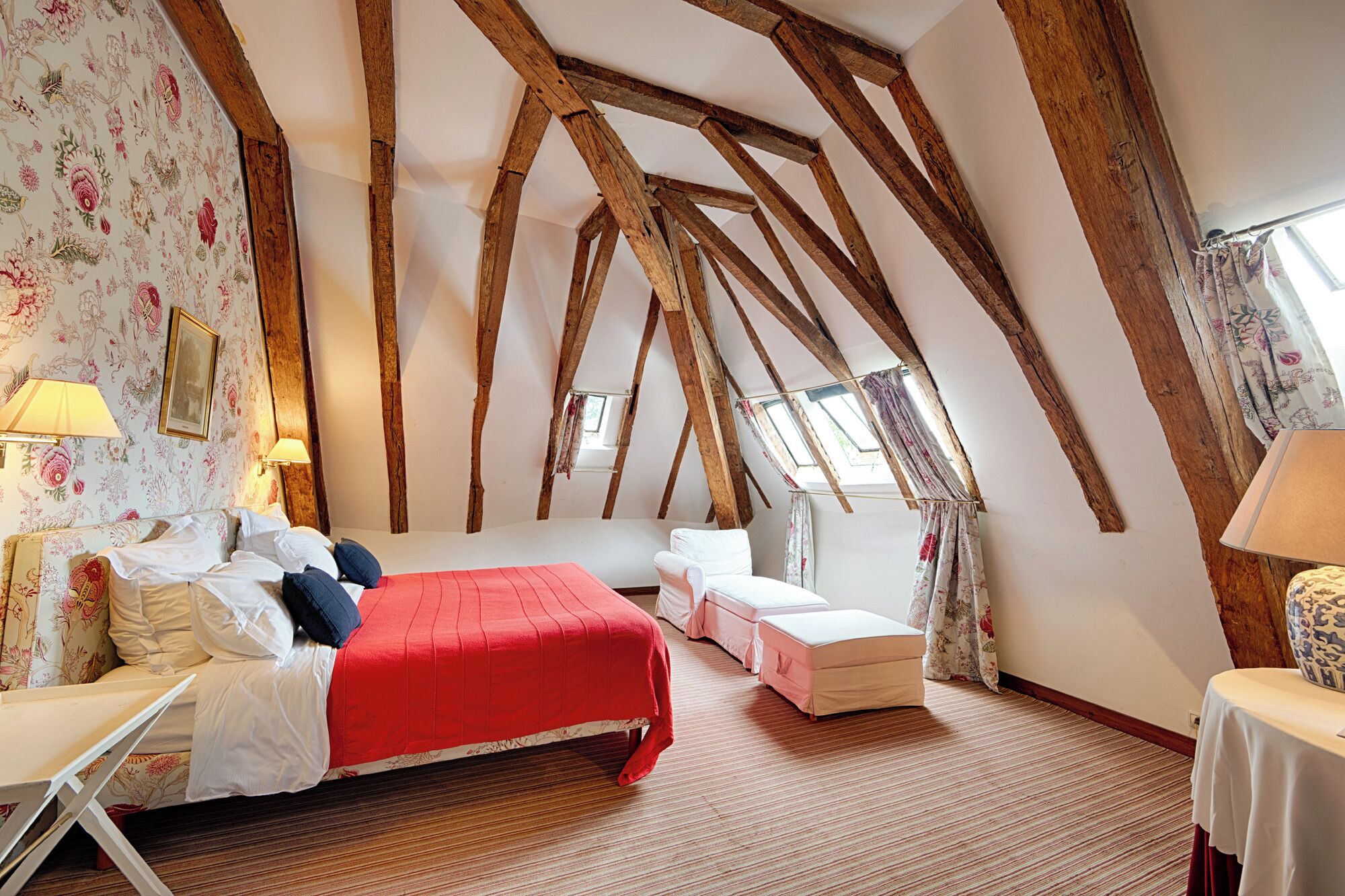 A bedroom with exposed wooden beams, a red bedspread, and a floral wallpaper.