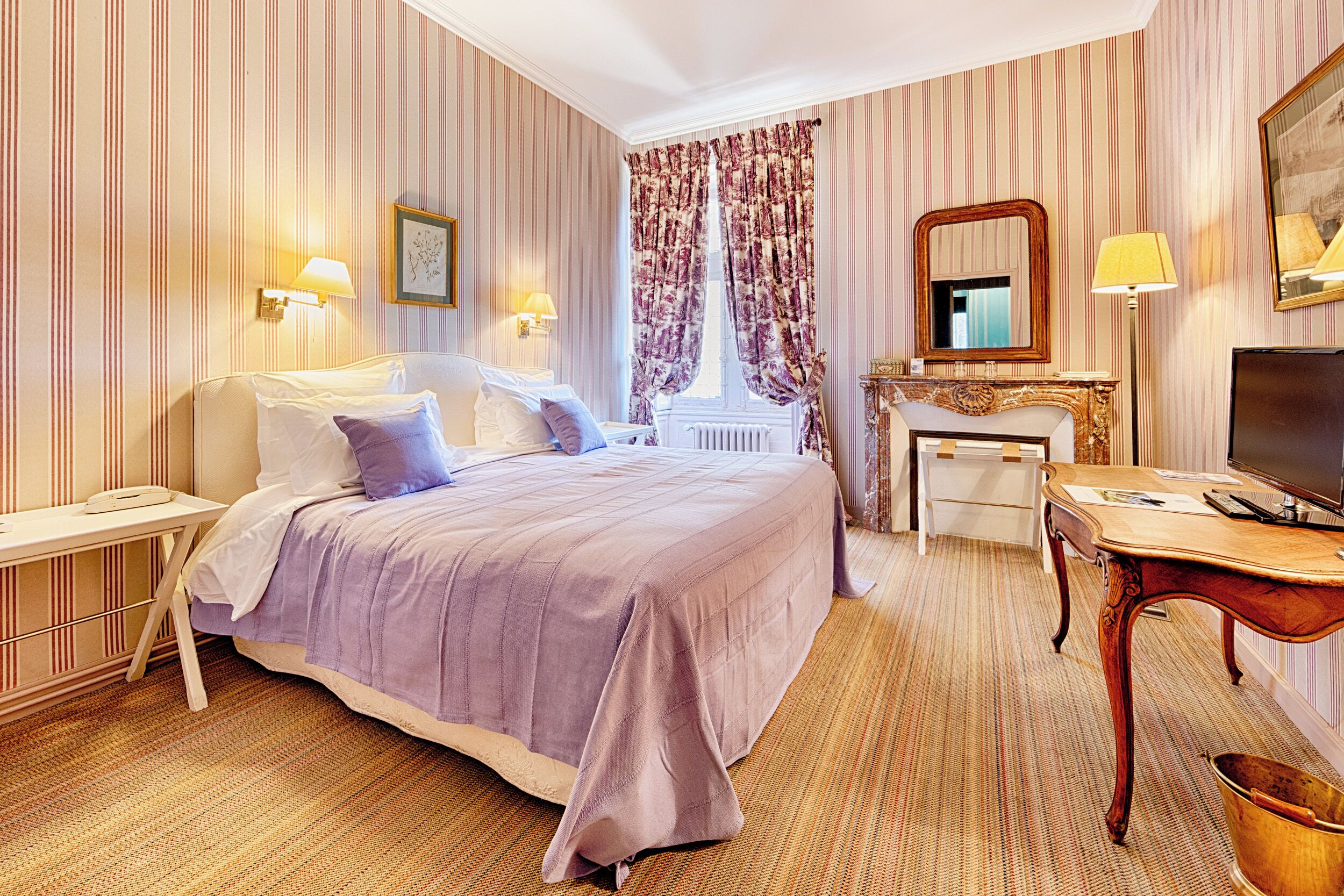 A hotel room with a bed, bedside tables, a desk, a television, and a window with curtains.