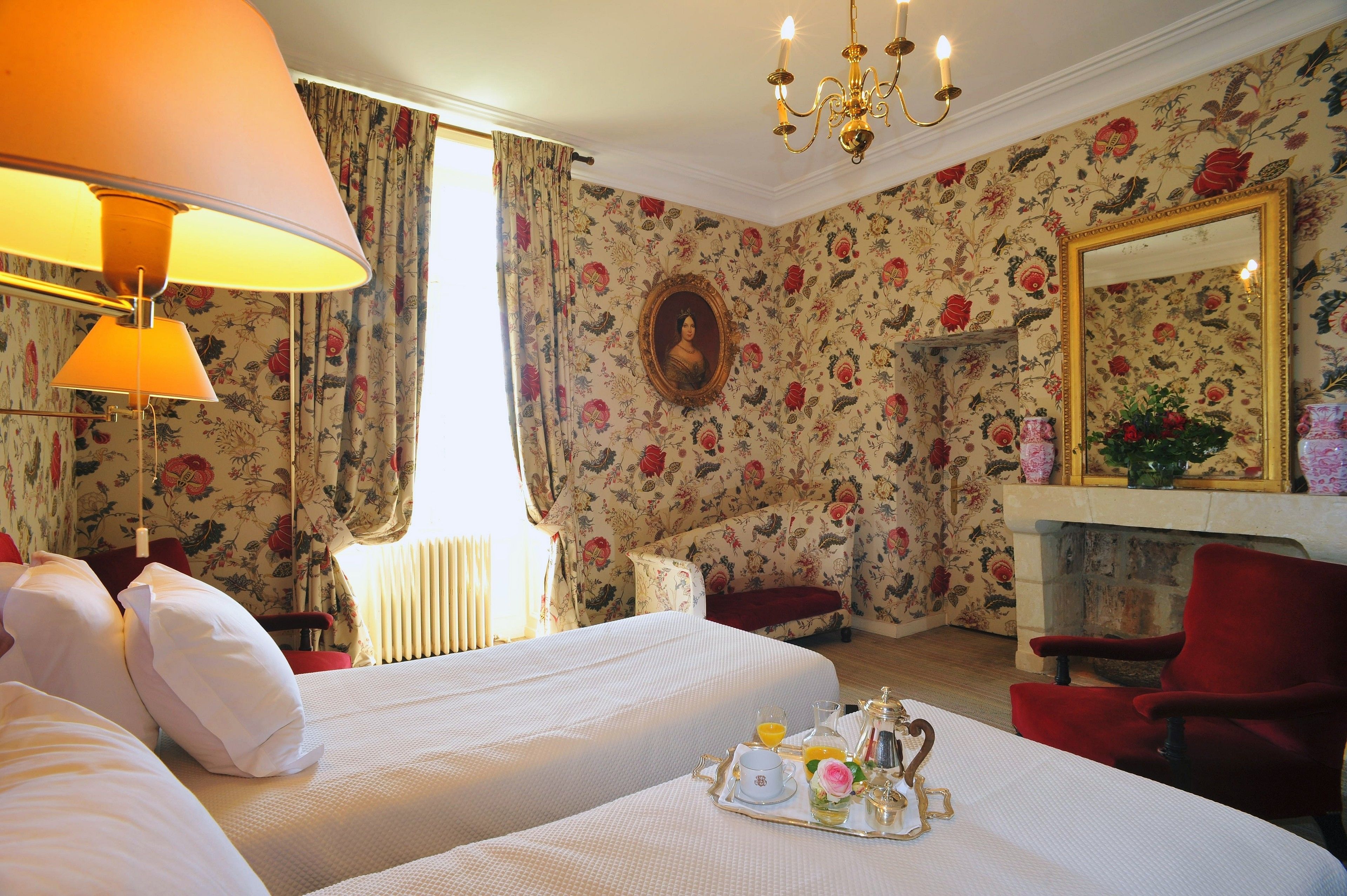 A hotel room with floral wallpaper, a chandelier, a fireplace, and a bed with a tray of breakfast items.