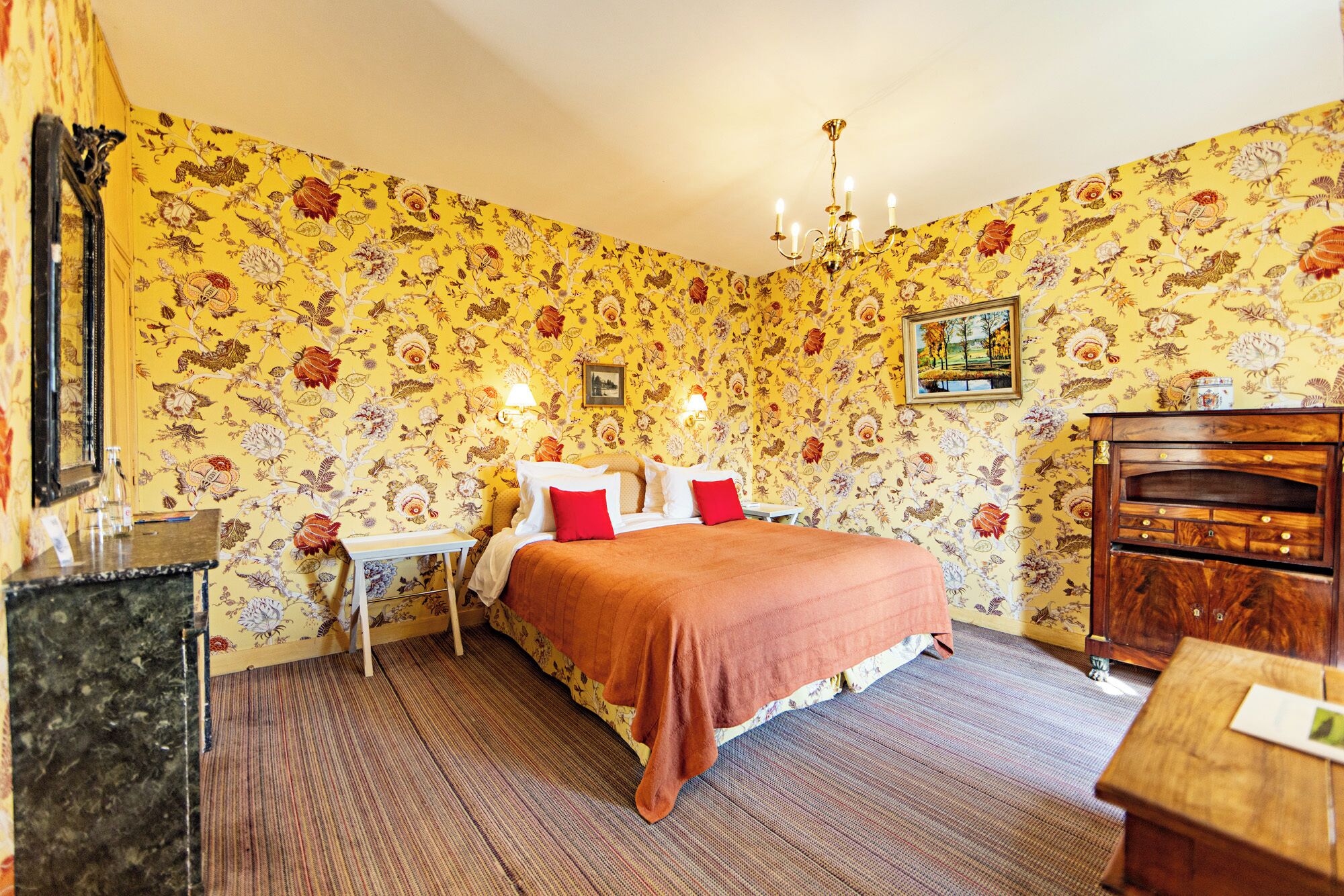 A room with floral wallpaper, a bed with red and white bedding, a wooden dresser, and a chandelier.