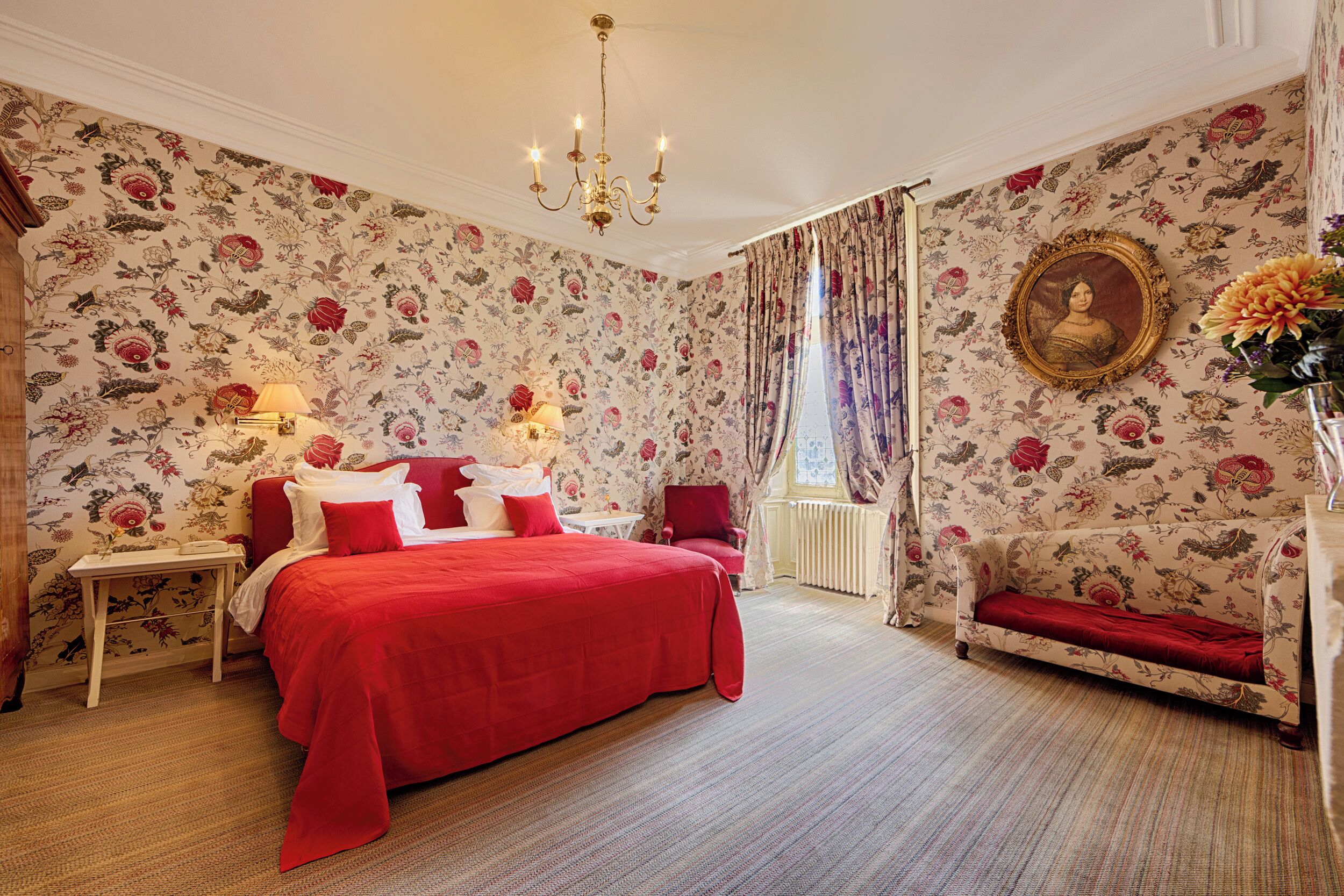 A bedroom with floral wallpaper, a red bed, a red armchair, a nightstand, a lamp, and a framed portrait.