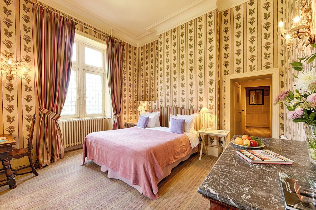 A bedroom with a large bed, a bedside table, a nightstand, a window with curtains, and a view of the hallway.