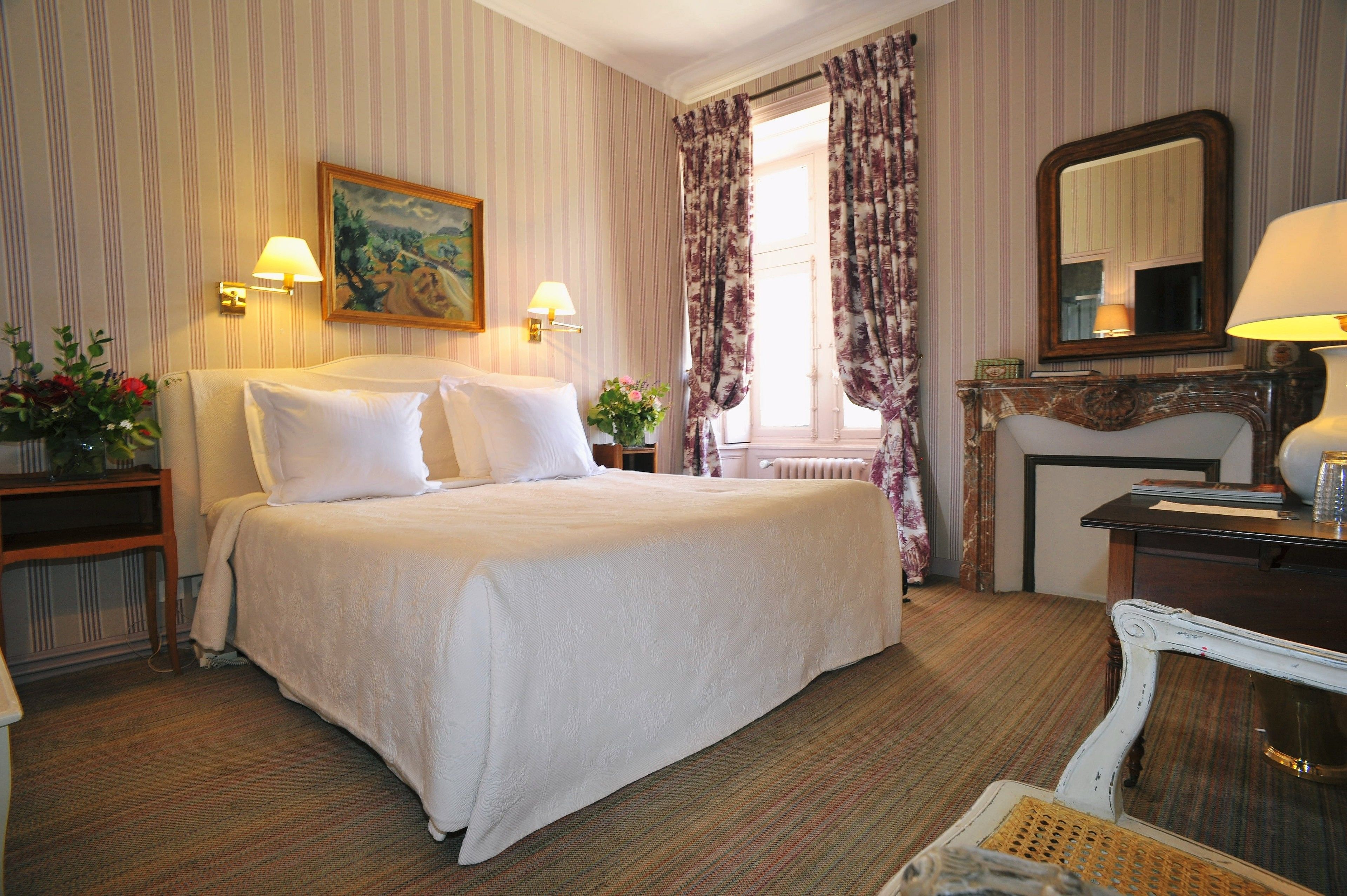 Une chambre d’hôtel avec un grand lit, deux lampes de chevet, un tableau au mur, une fenêtre avec des rideaux, une commode et un miroir.