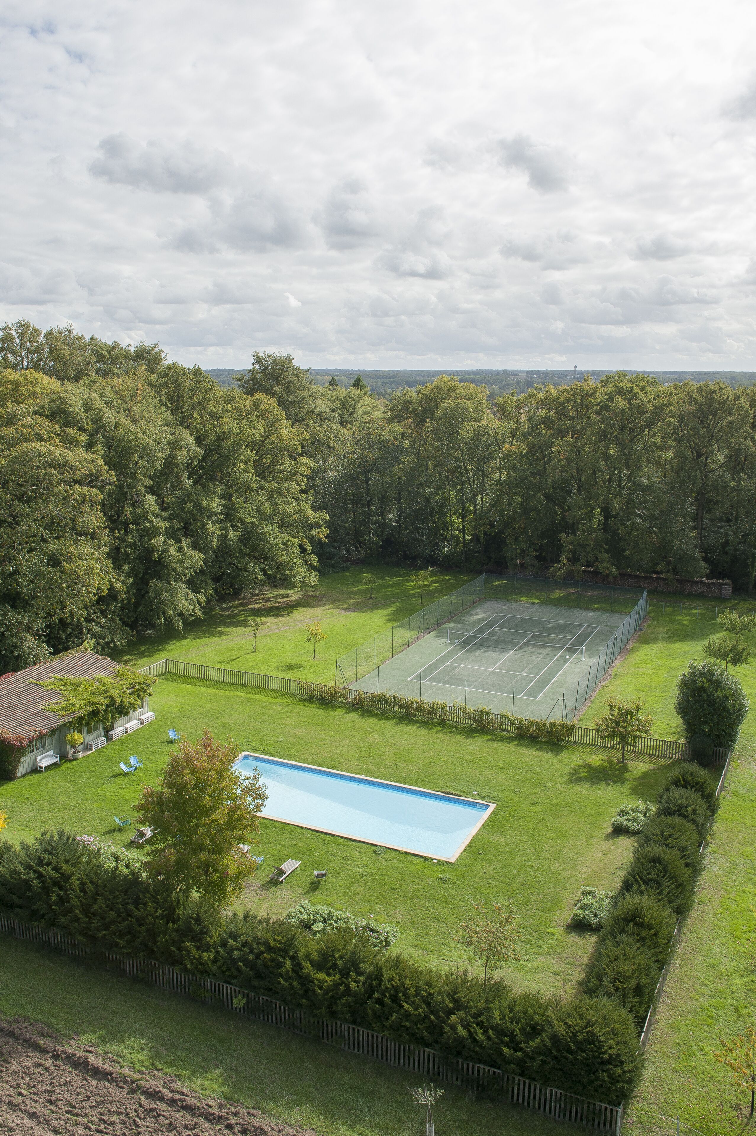 A tennis court surrounded by a fence, a swimming pool, and a grassy area with trees.