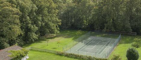 A tennis court surrounded by a fence, a swimming pool, and a grassy area with trees.