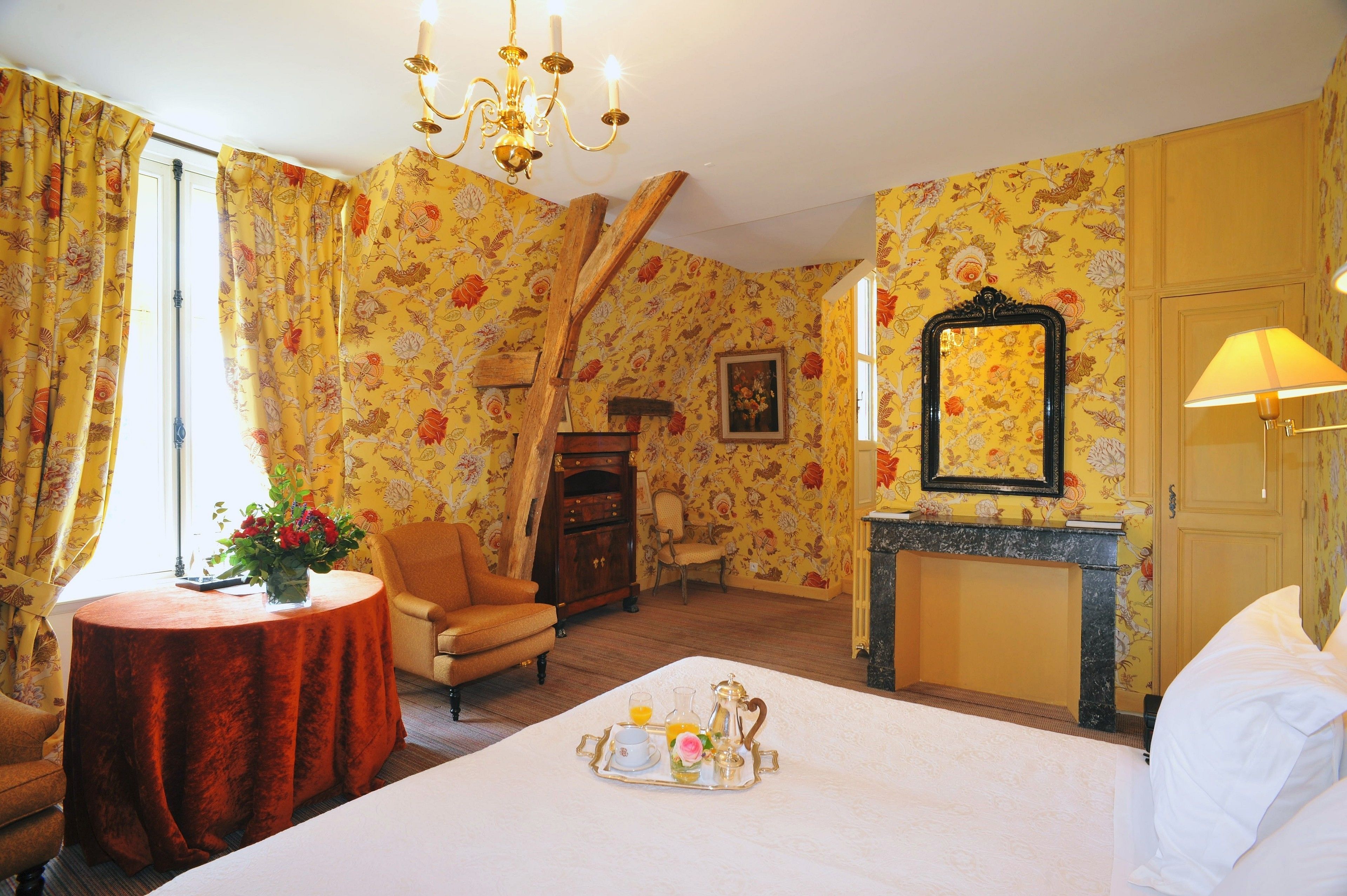 A hotel room with floral wallpaper, a bed with a tray of breakfast, a fireplace, and a chandelier.