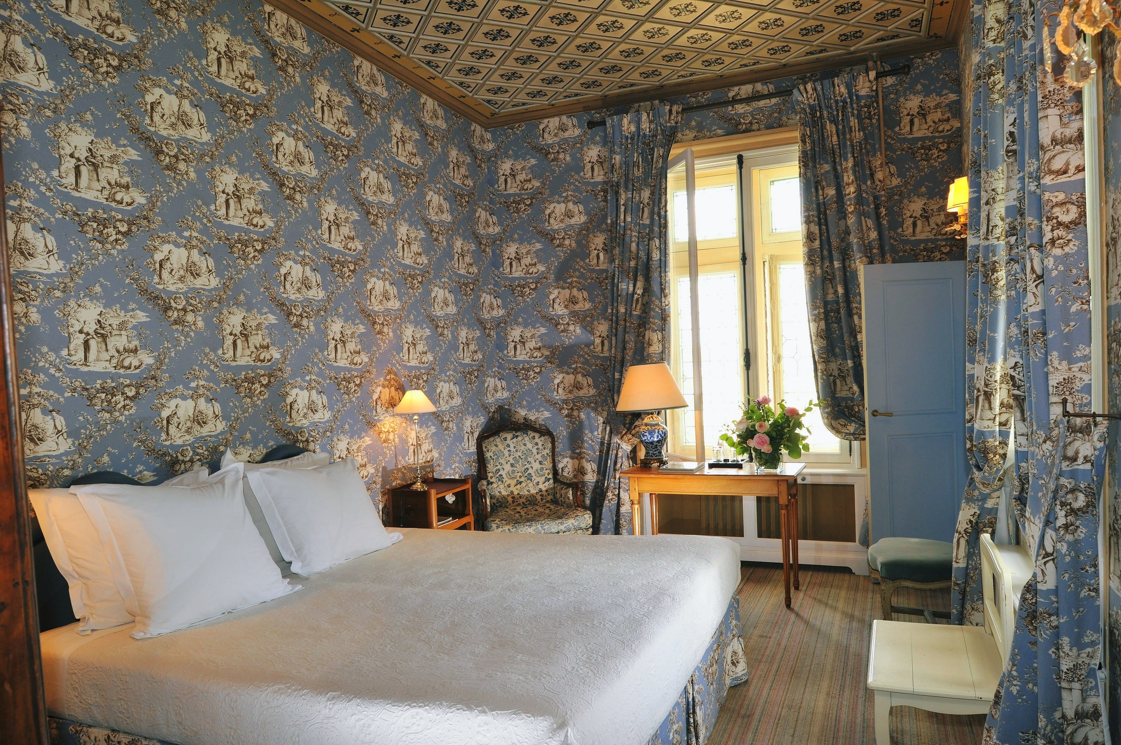 A bedroom with a large bed, two bedside tables with lamps, a chair, a small table with a vase of flowers, and a window with curtains.