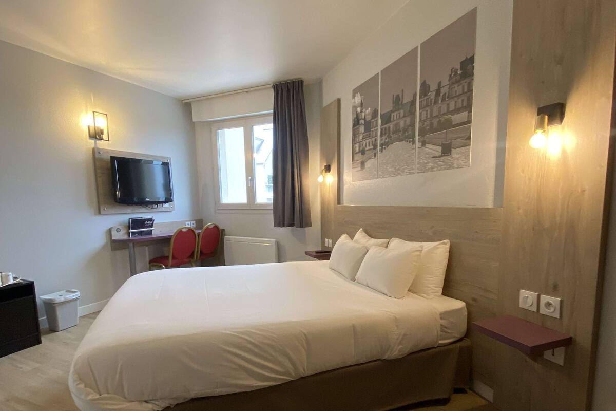 A modern hotel room with a large bed, a flat-screen TV mounted on the wall, a desk with a chair, and a window with curtains.